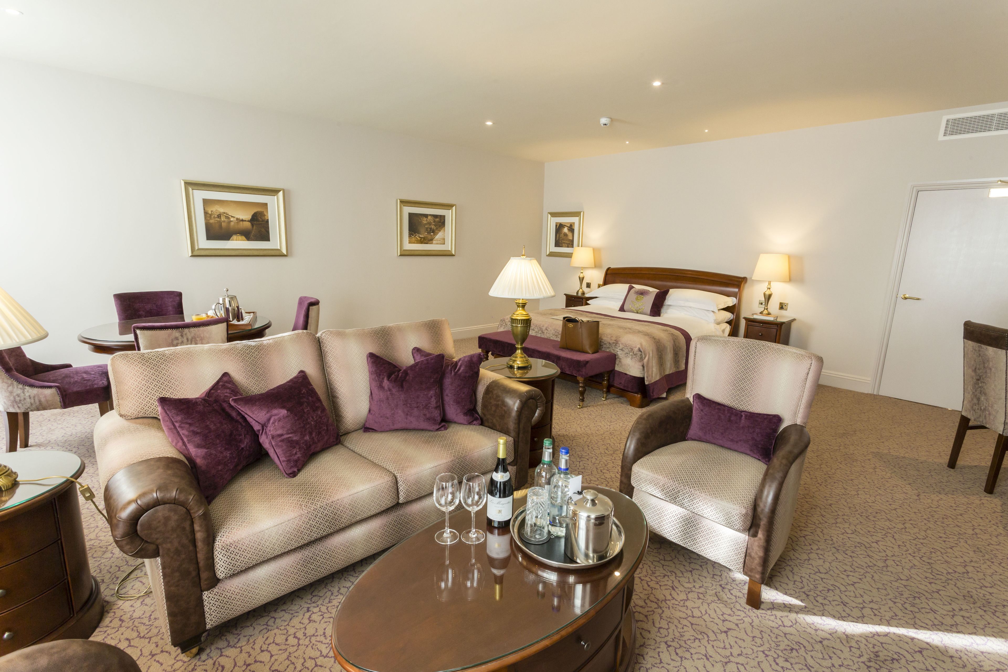 A hotel room with a sofa set, a coffee table with wine glasses and bottles, a bed with a headboard, and two bedside tables with lamps.