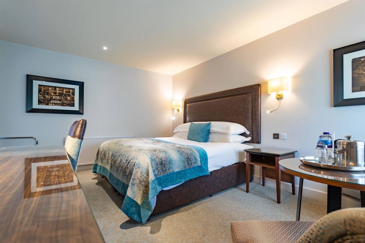 Een hotelkamer met een bed, nachtkastje, stoel en een ingelijst schilderij aan de muur.