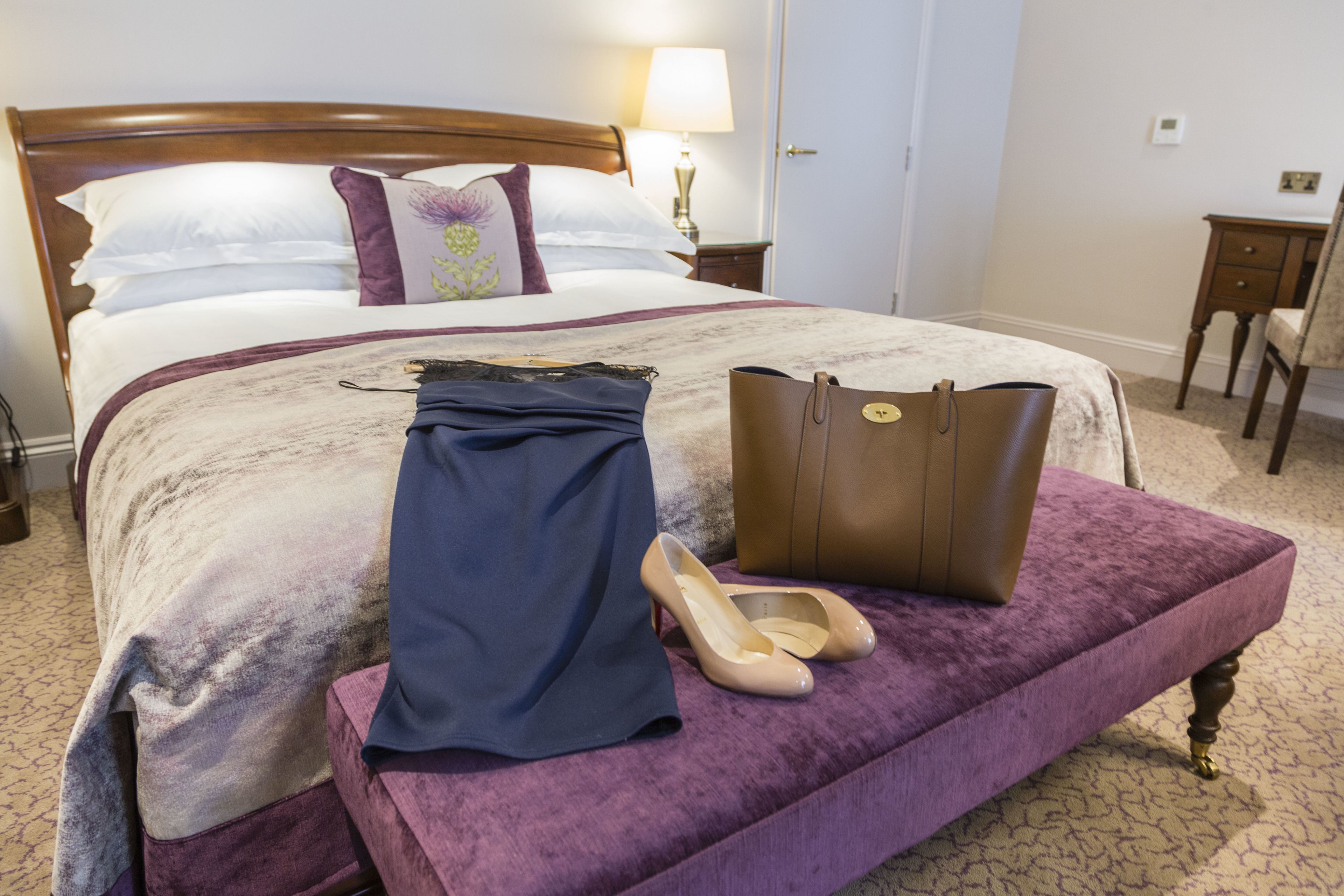 Ein ordentlich bezogenes Bett mit einer violetten Decke, einem Paar High Heels und einer braunen Handtasche auf einem passenden Pouf.