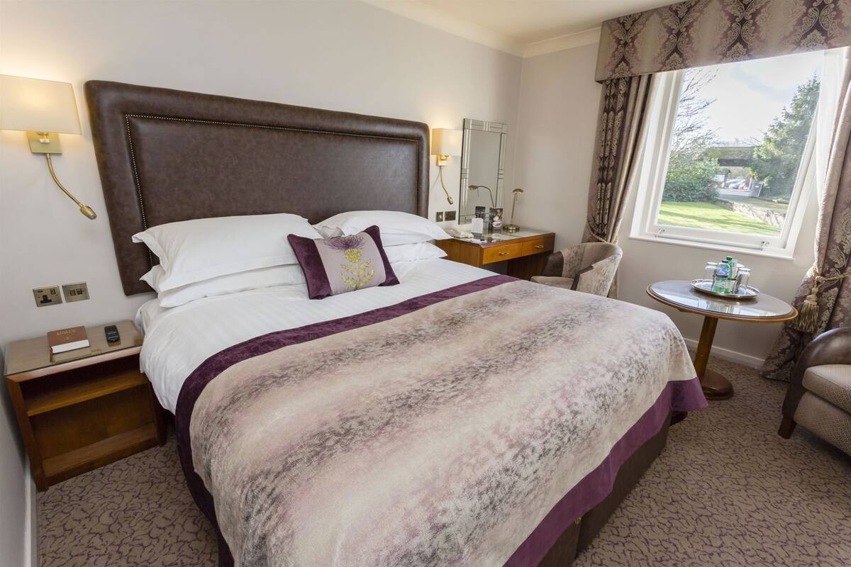 Een hotelkamer met een groot bed, nachtkastje, stoel en een raam met gordijnen, met uitzicht op de omgeving.