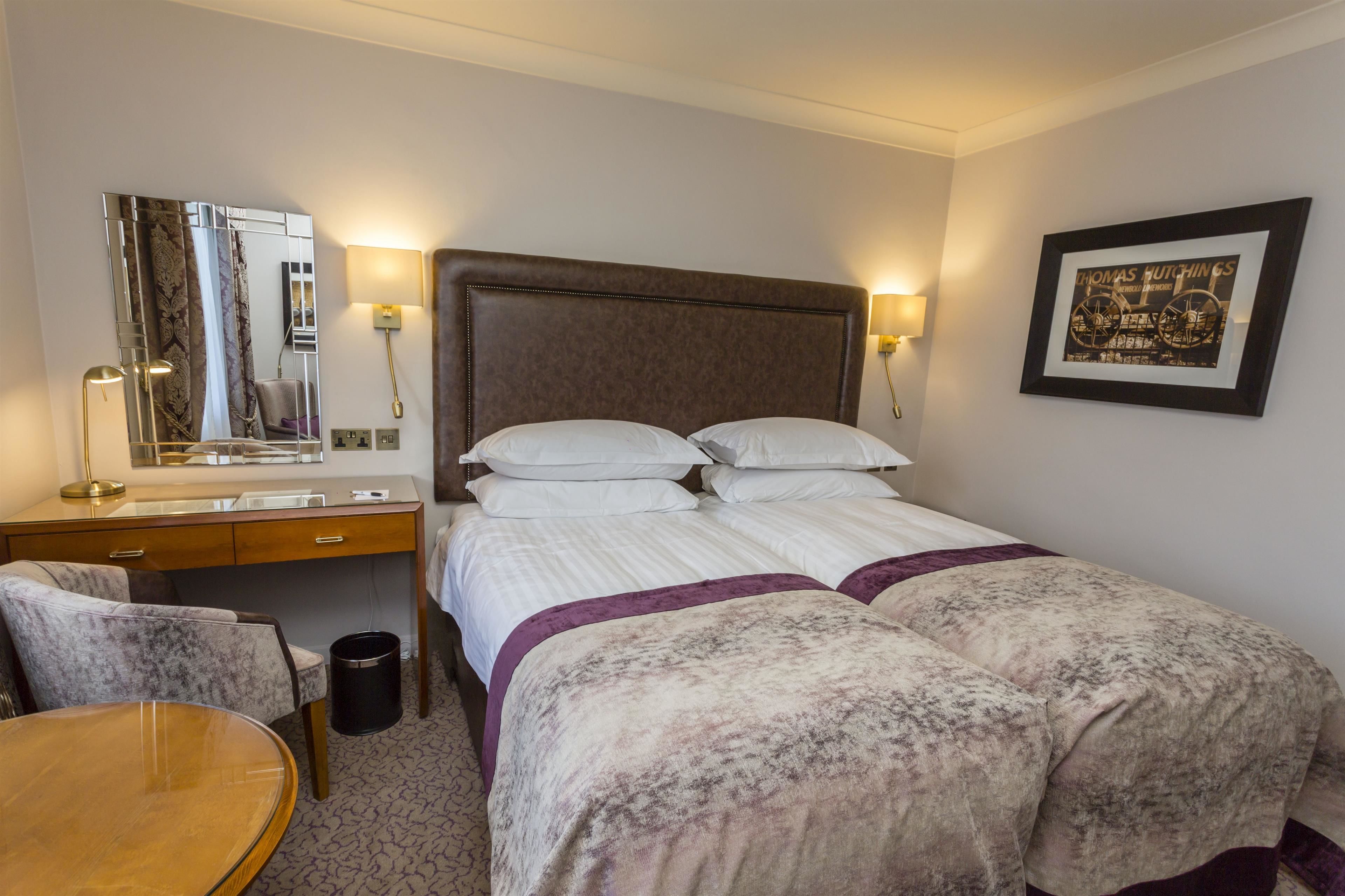 A hotel room with a bed, a desk with a mirror, a chair, and a framed picture on the wall.