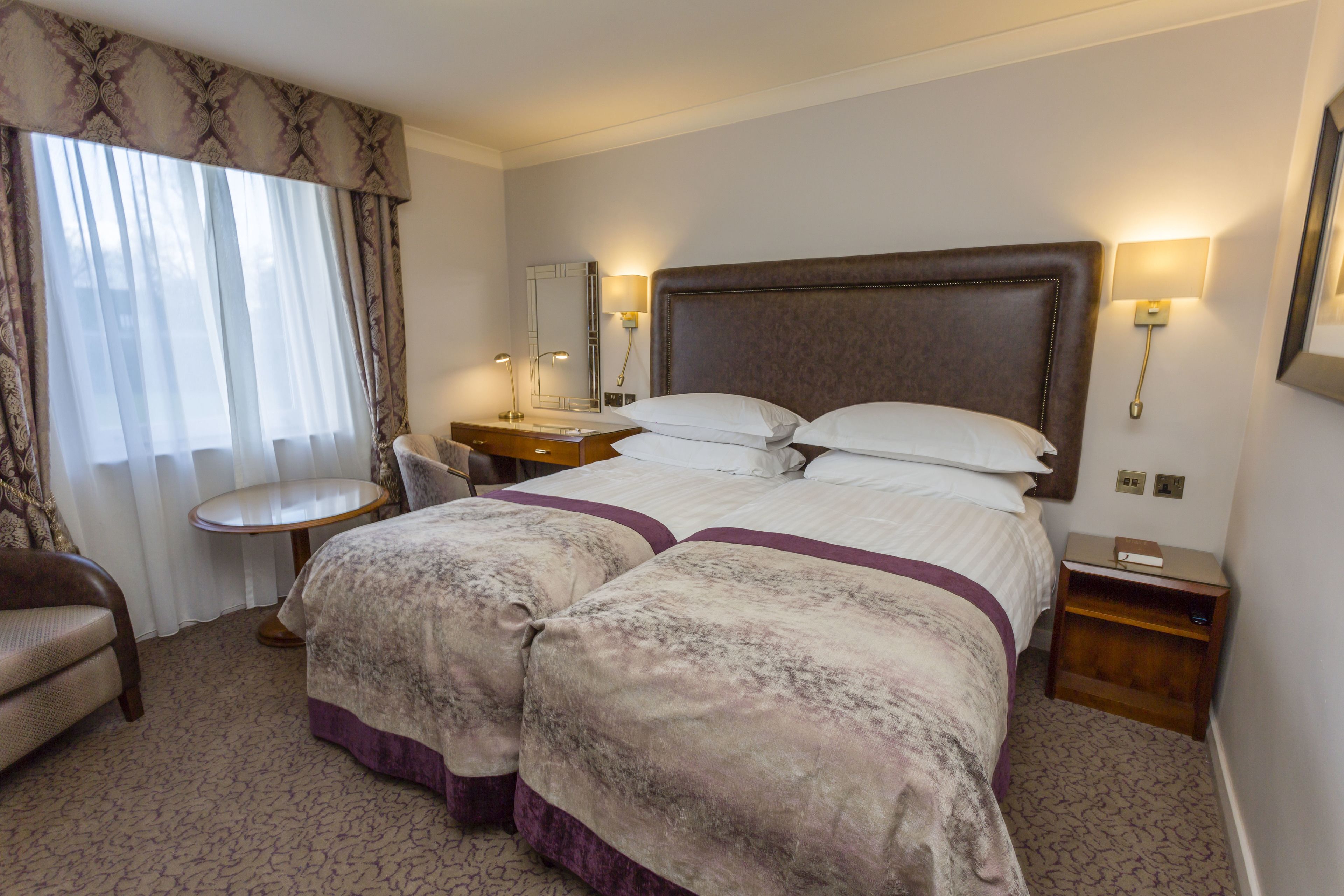 A hotel room with a large bed, two bedside tables, a chair, a small table, and a window with curtains.
