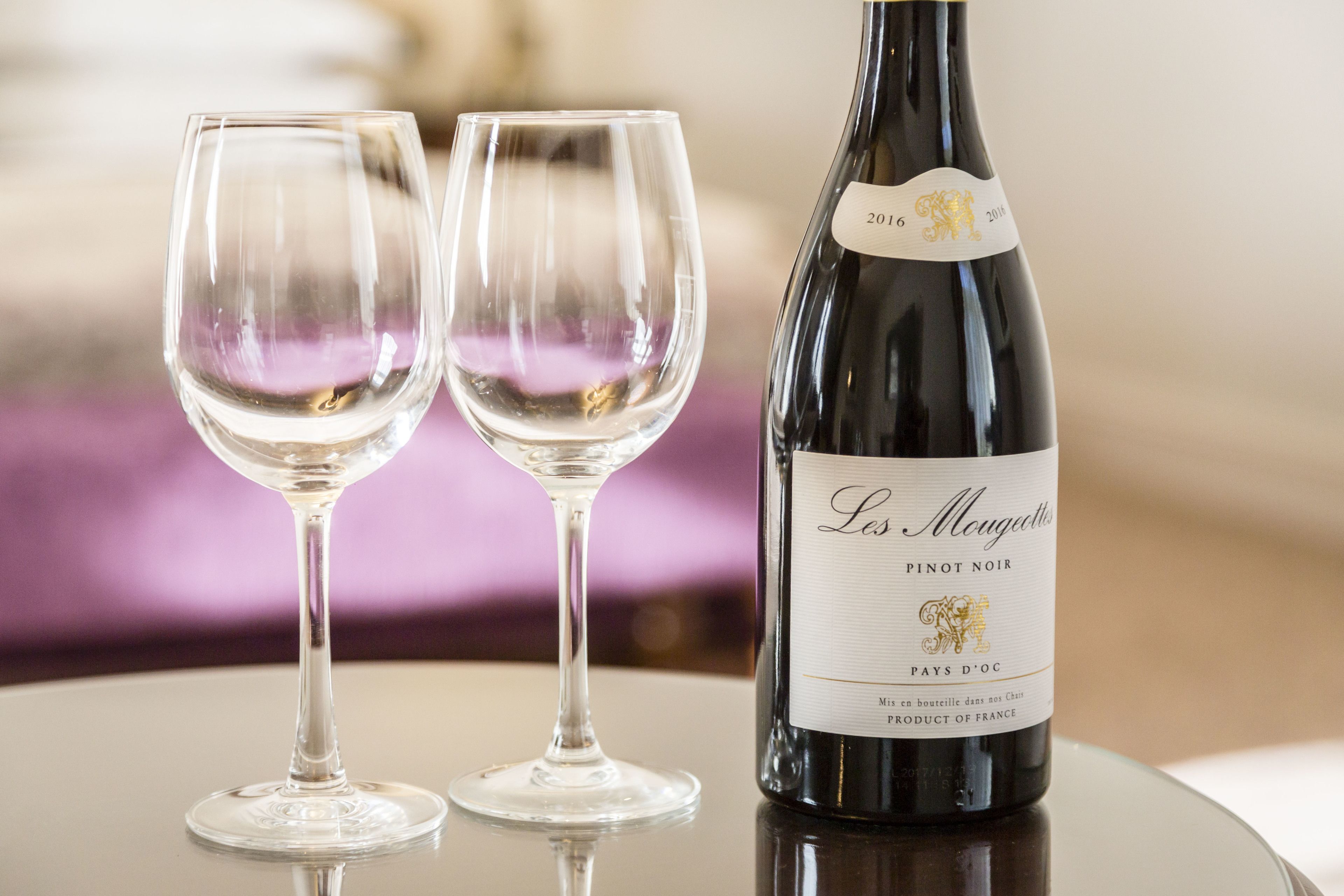 A bottle of Les Maugédottes Pinot Noir wine and two empty wine glasses on a reflective surface.