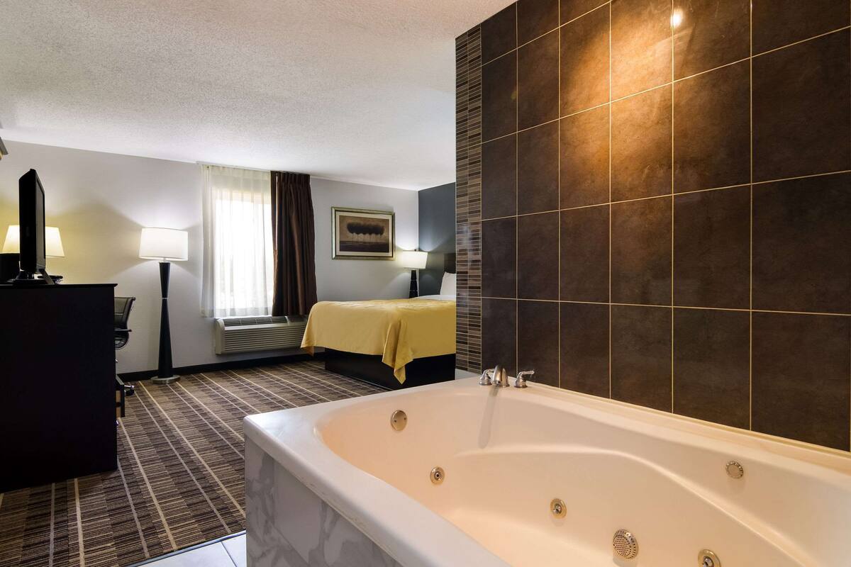 A hotel room with a large bathtub, a bed with a yellow bedspread, a desk with a lamp, and a window with curtains.