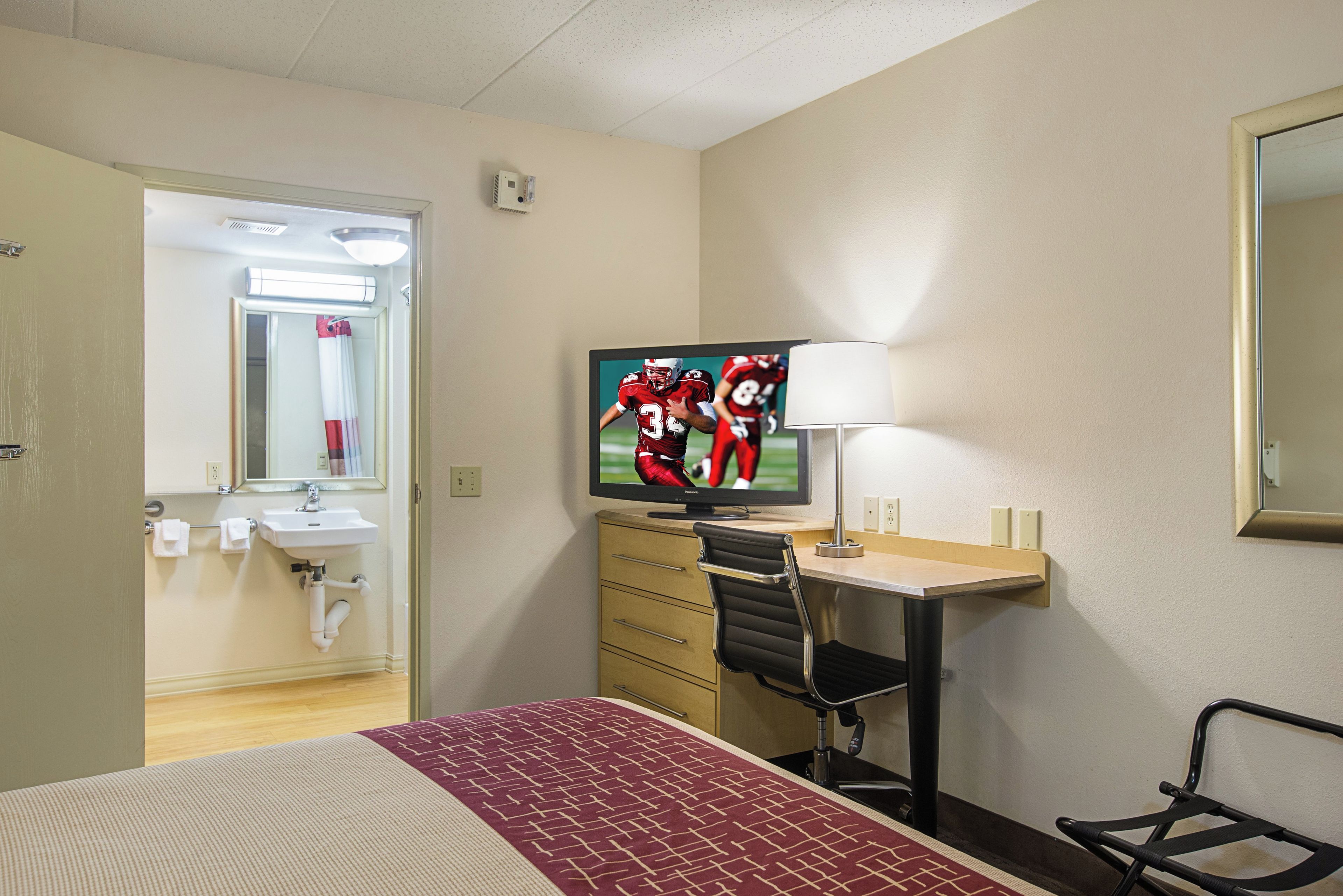 A hotel room with a bed, a desk with a chair, a television, a mirror, and a bathroom with a sink and towel rack.