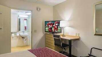 A hotel room with a bed, a desk with a chair, a television, a mirror, and a bathroom with a sink and towel rack.