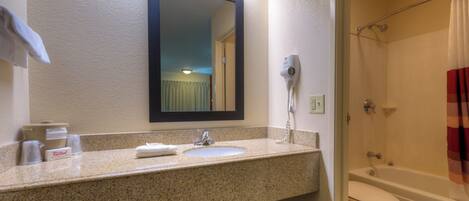 A bathroom with a granite countertop, a sink, a mirror, a hairdryer, and a shower with a curtain.