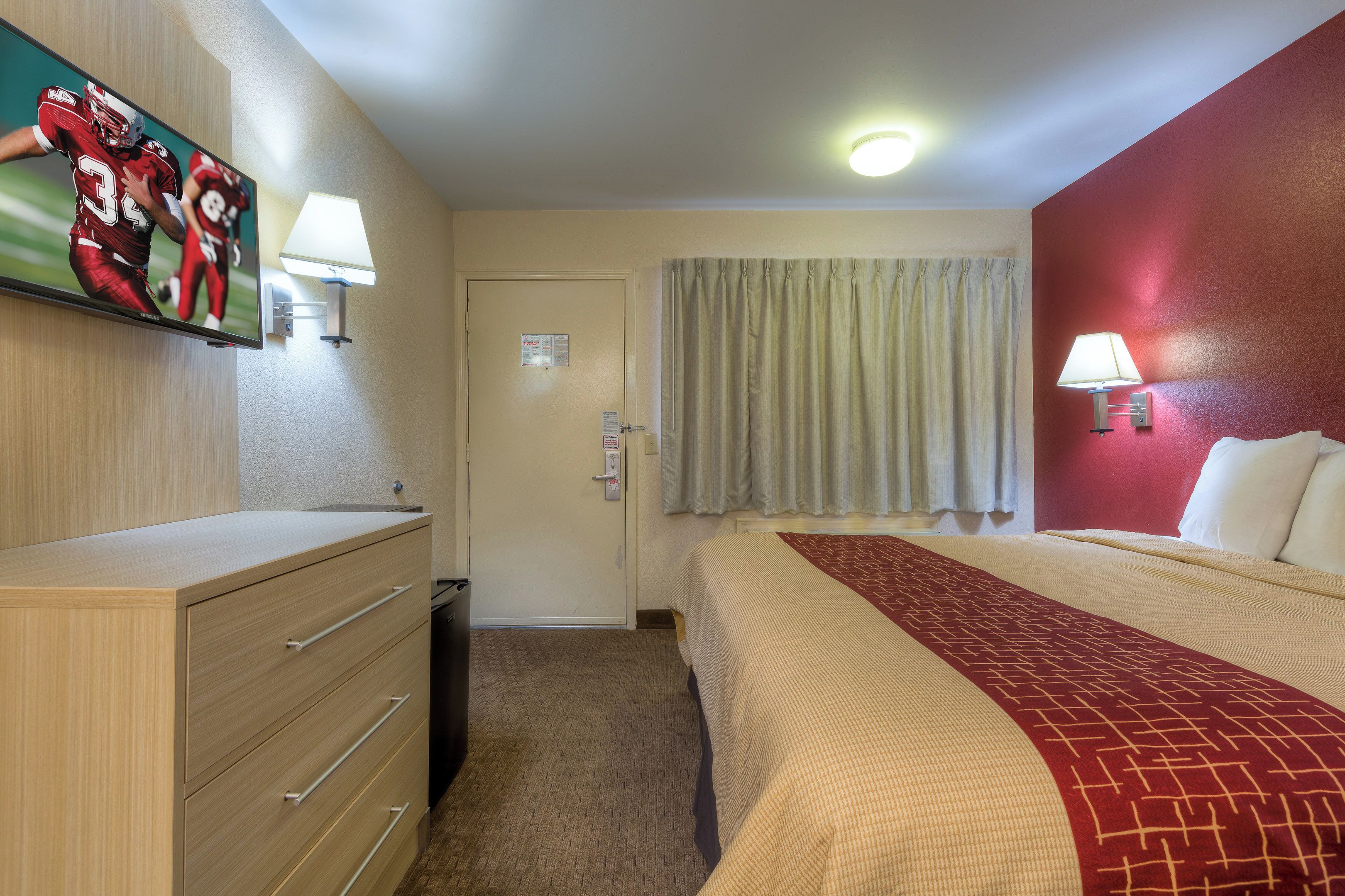 A hotel room with a bed, a dresser, a television mounted on the wall, and a window with curtains.