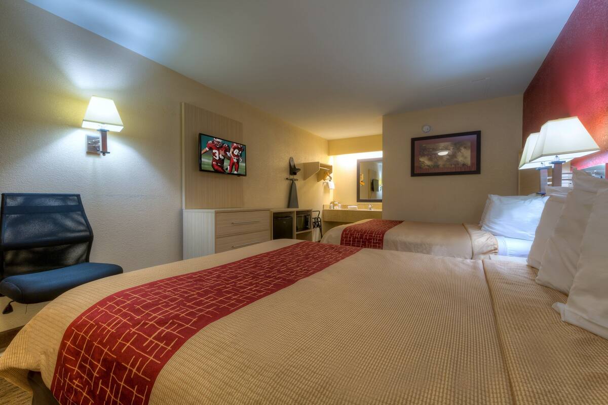 A hotel room with a large bed, a desk, a chair, a desk lamp, a wall-mounted TV, and a painting.