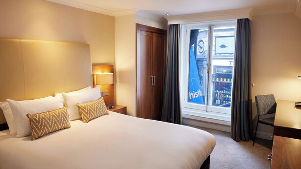 A hotel room with a large bed, a nightstand with a lamp, a wardrobe, a window with curtains, and a chair. - Thistle London Piccadilly (London)