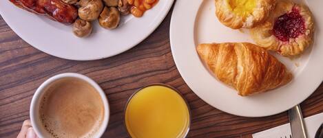 A breakfast setting with plates of eggs, sausages, beans, tomatoes, and croissants, along with cups of coffee and juice.