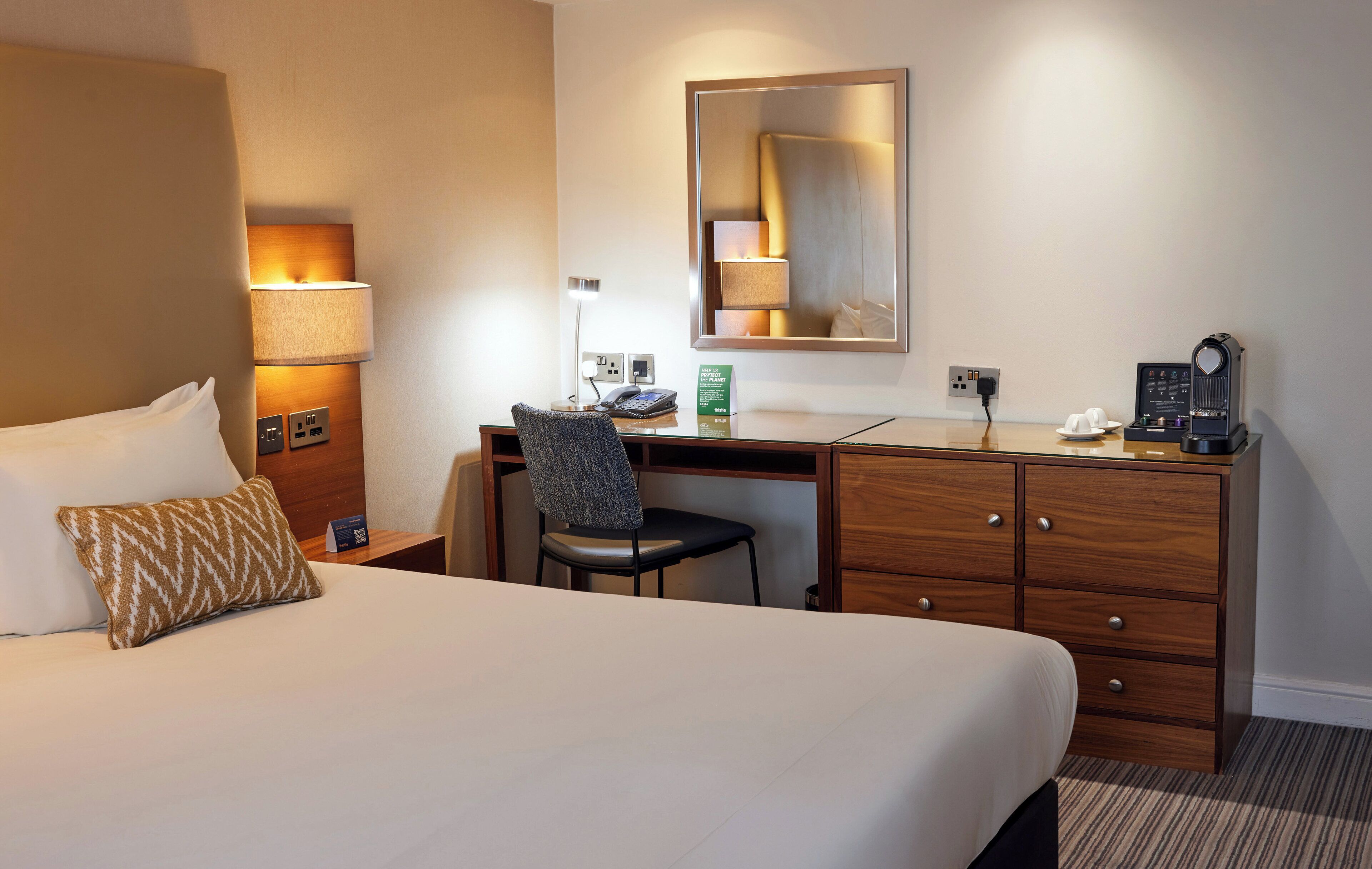 A hotel room with a large bed, a desk with a chair, a dresser, a lamp, a coffee maker, and a mirror.