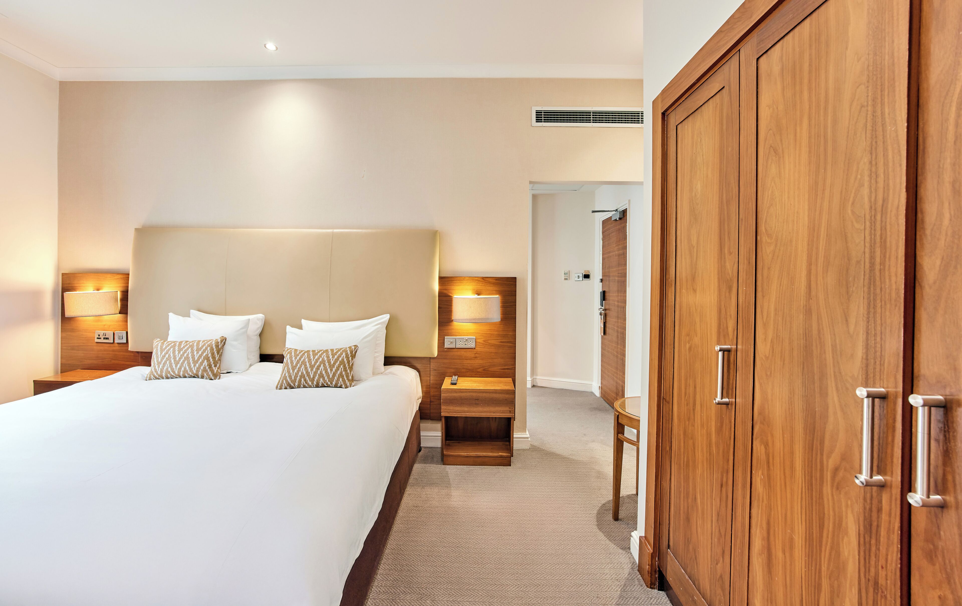 Une chambre d’hôtel avec un grand lit, deux tables de chevet avec des lampes, une armoire et une porte donnant sur une autre pièce.