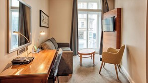 A hotel room with a wooden desk, a sofa, a round table, a chair, and a TV mounted on the wall. - Thistle London Piccadilly (London)
