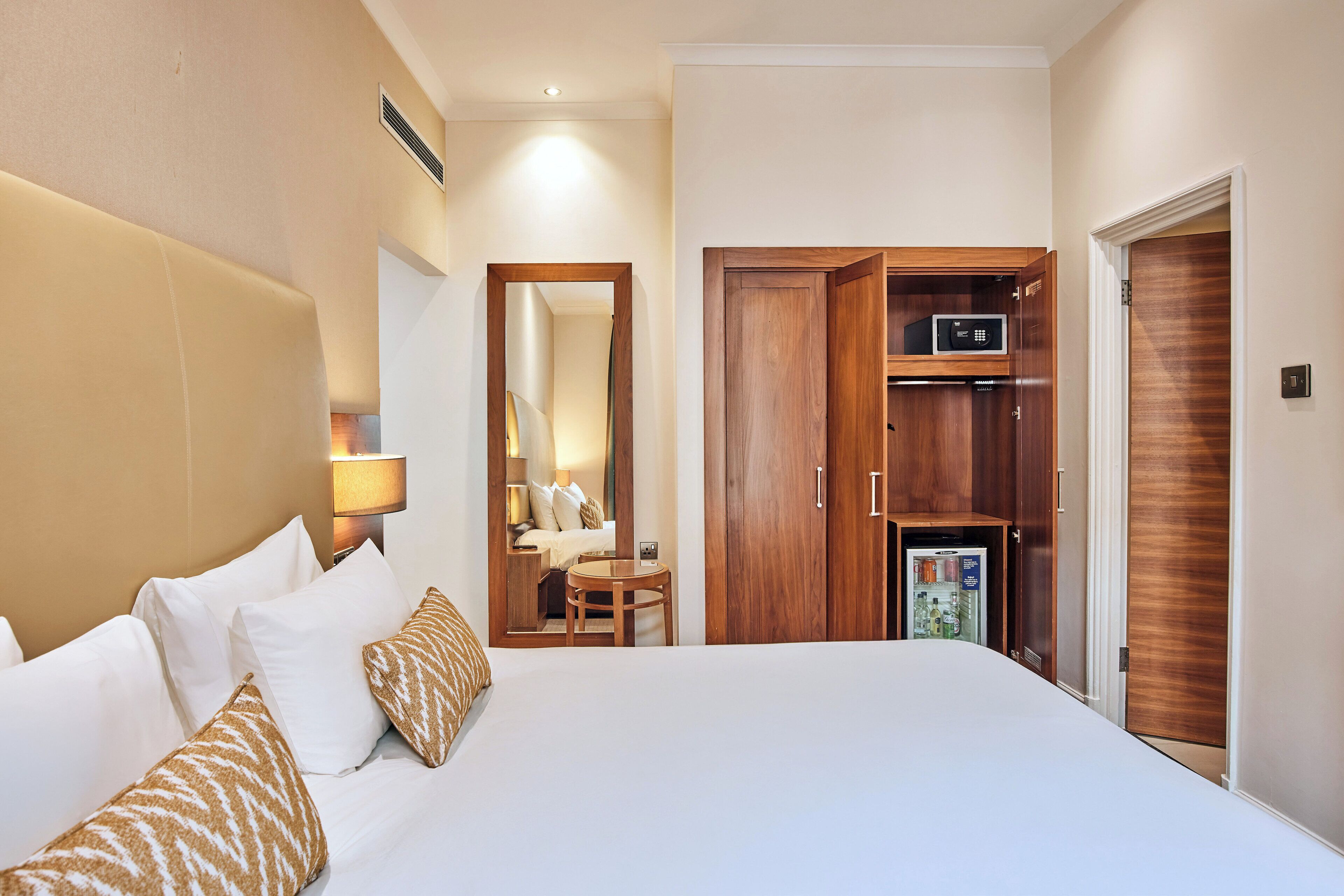 A hotel room with a large bed, a bedside table, a mirror, a wardrobe, and a minibar.