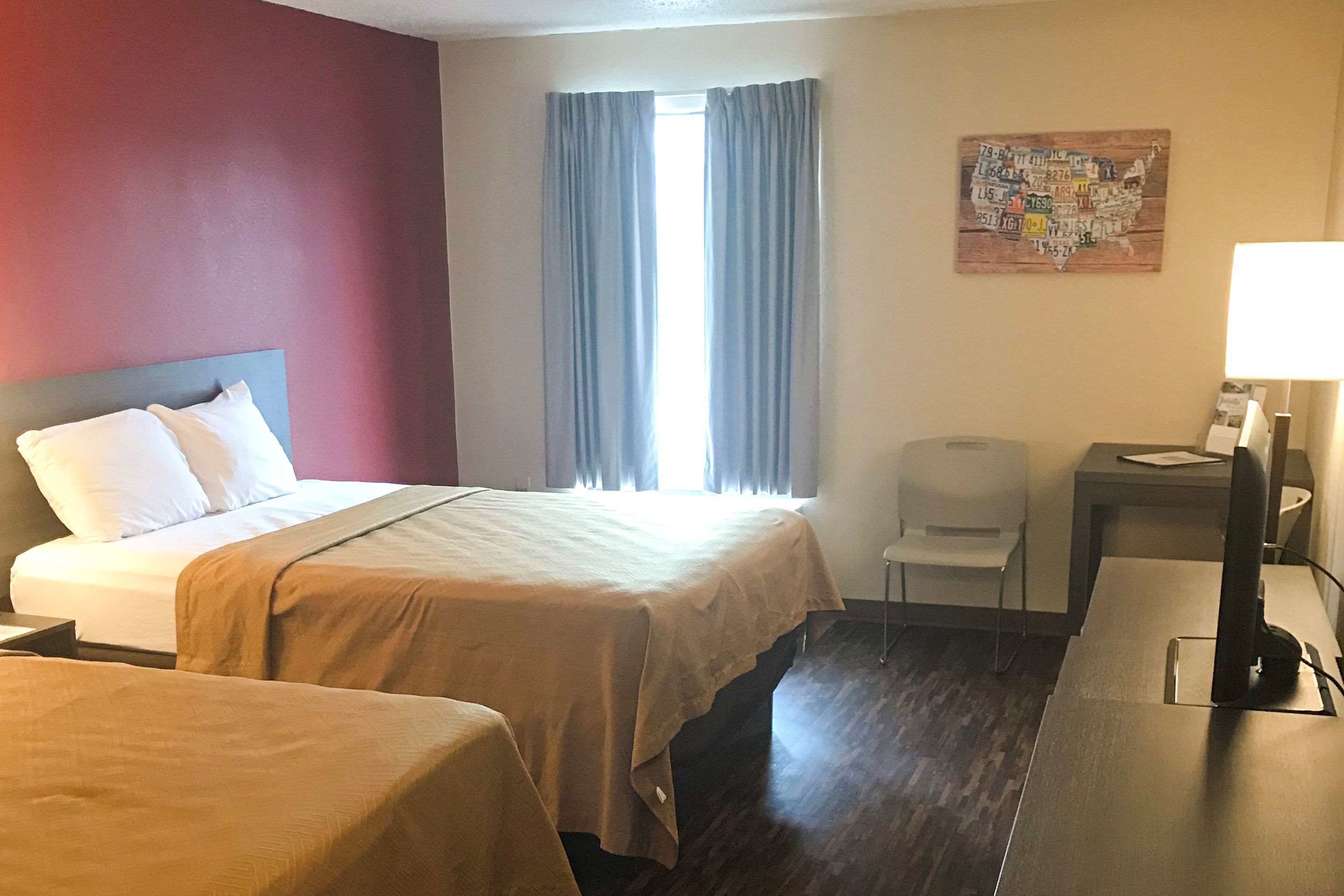 A hotel room with two beds, a desk with a lamp, a chair, a map on the wall, and a window with curtains.