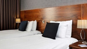 A hotel room with a wooden headboard, two beds with white linens, a nightstand with a lamp, and a patterned wall.