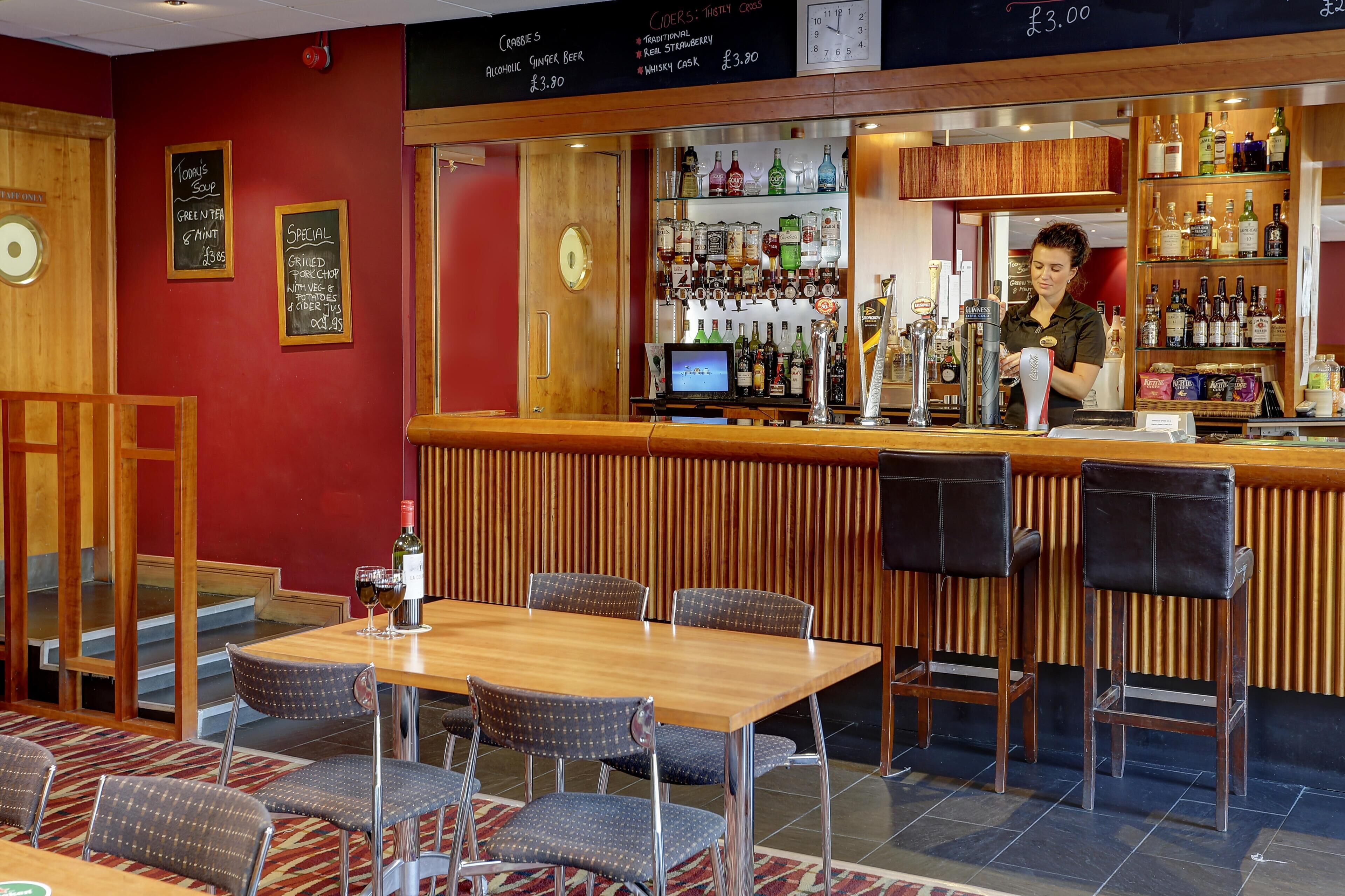 Una zona de bar con mostrador de madera, taburetes y un área de estar con mesas y sillas.