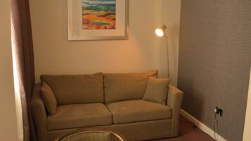 A room with a brown sofa, a wooden chair, a table with a floral tablecloth, a framed painting, a floor lamp, and a patterned wall.