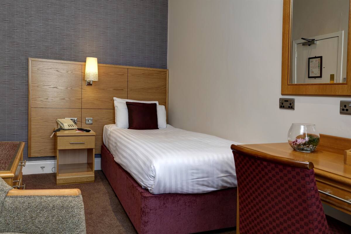 A hotel room with a wooden headboard, a bedside table with a lamp, a telephone, a bed with white linens and a dark pillow, a wooden desk, a chair, and a small vase with flowers.
