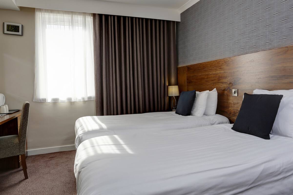 A hotel room with two beds, a desk with a chair, a lamp, and a window with curtains.
