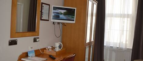 A hotel room with a wooden desk, a television mounted on the wall, a mirror, a cabinet, and a window with curtains.