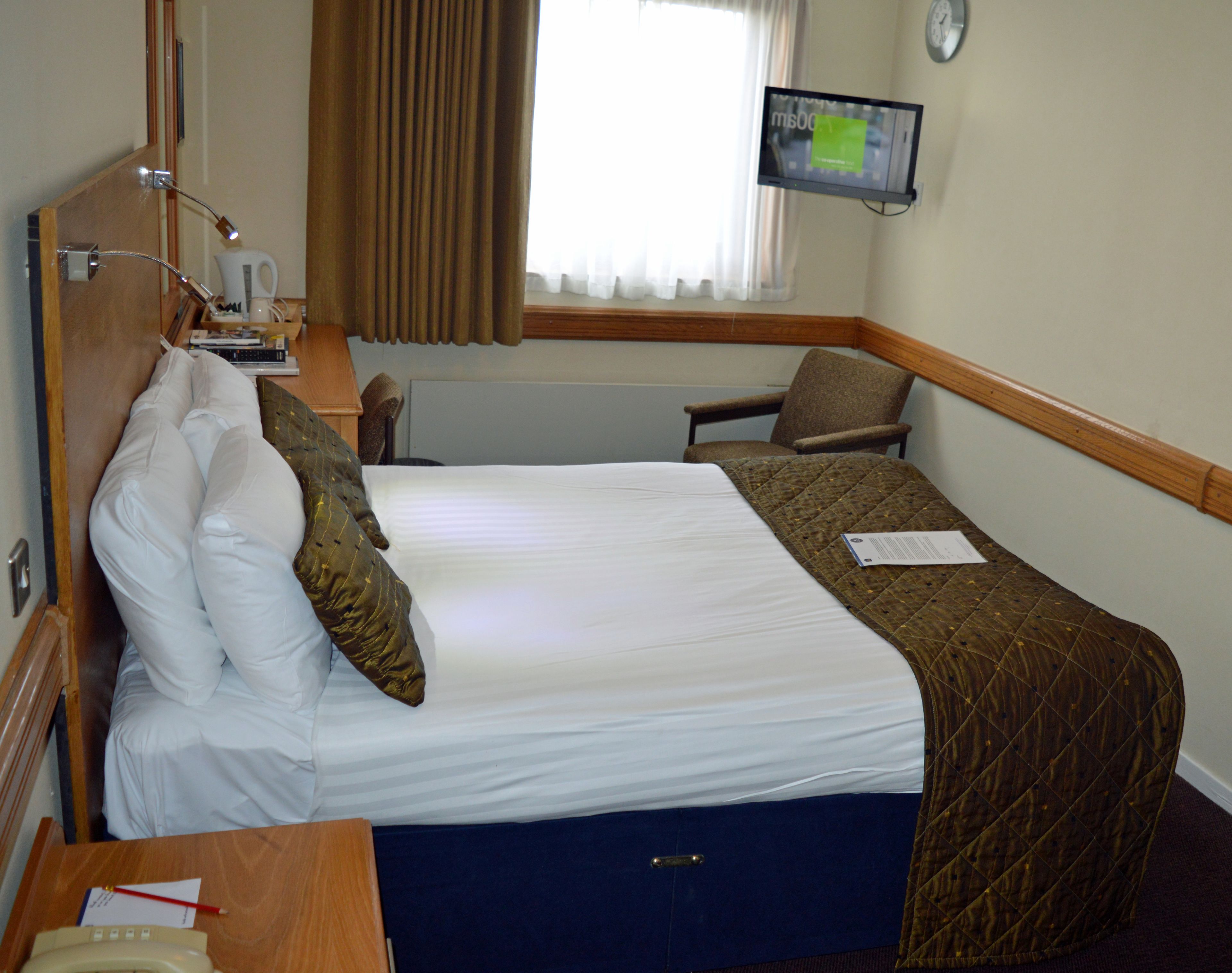 A hotel room with a bed, a desk with a telephone, a chair, a television mounted on the wall, and a window with curtains.