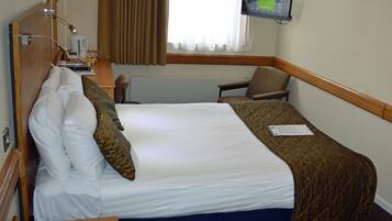 A hotel room with a bed, a desk with a telephone, a chair, a television mounted on the wall, and a window with curtains.