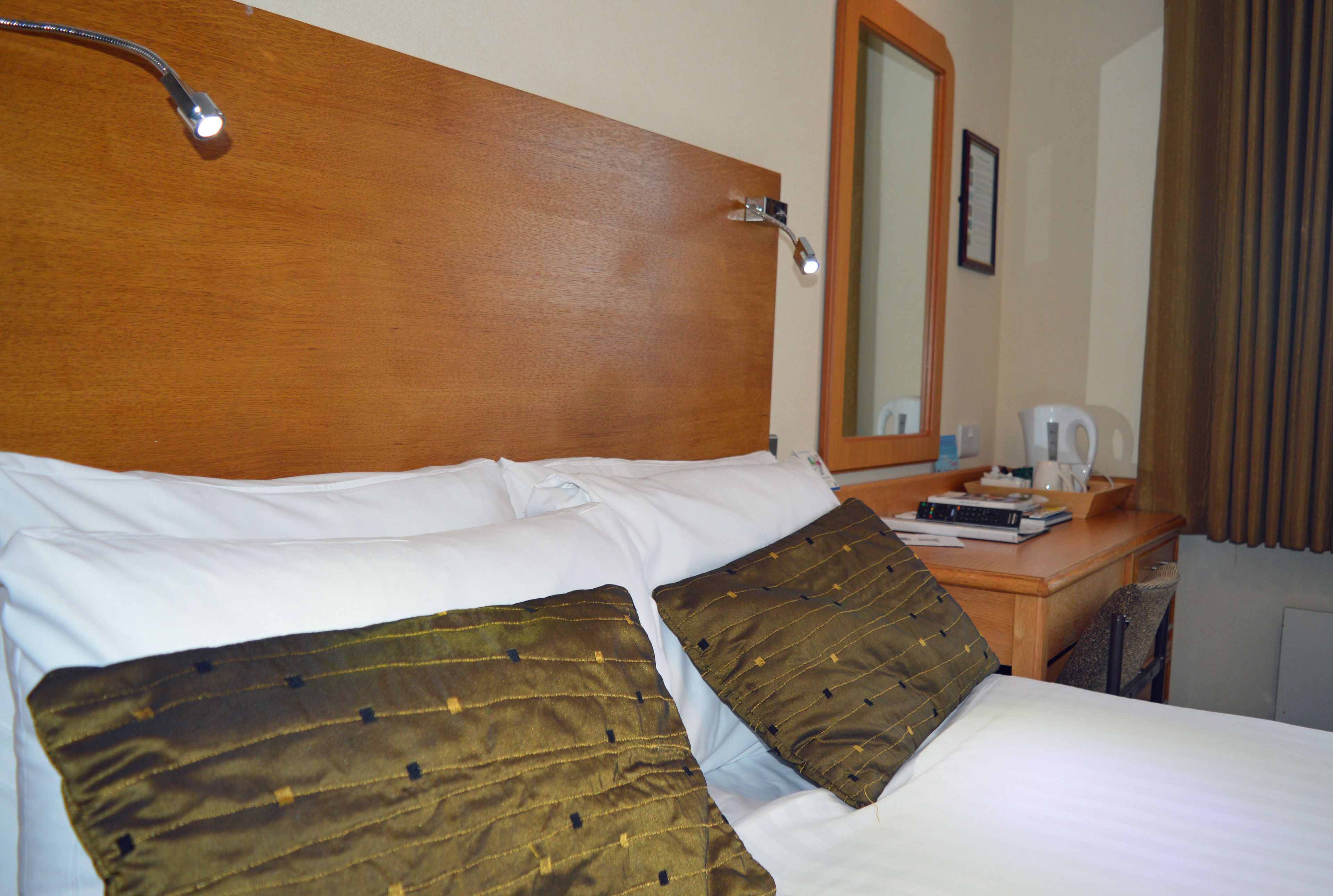 A neatly made bed with white and brown bedding, a wooden headboard, a bedside table with a lamp, and a mirror on the wall.
