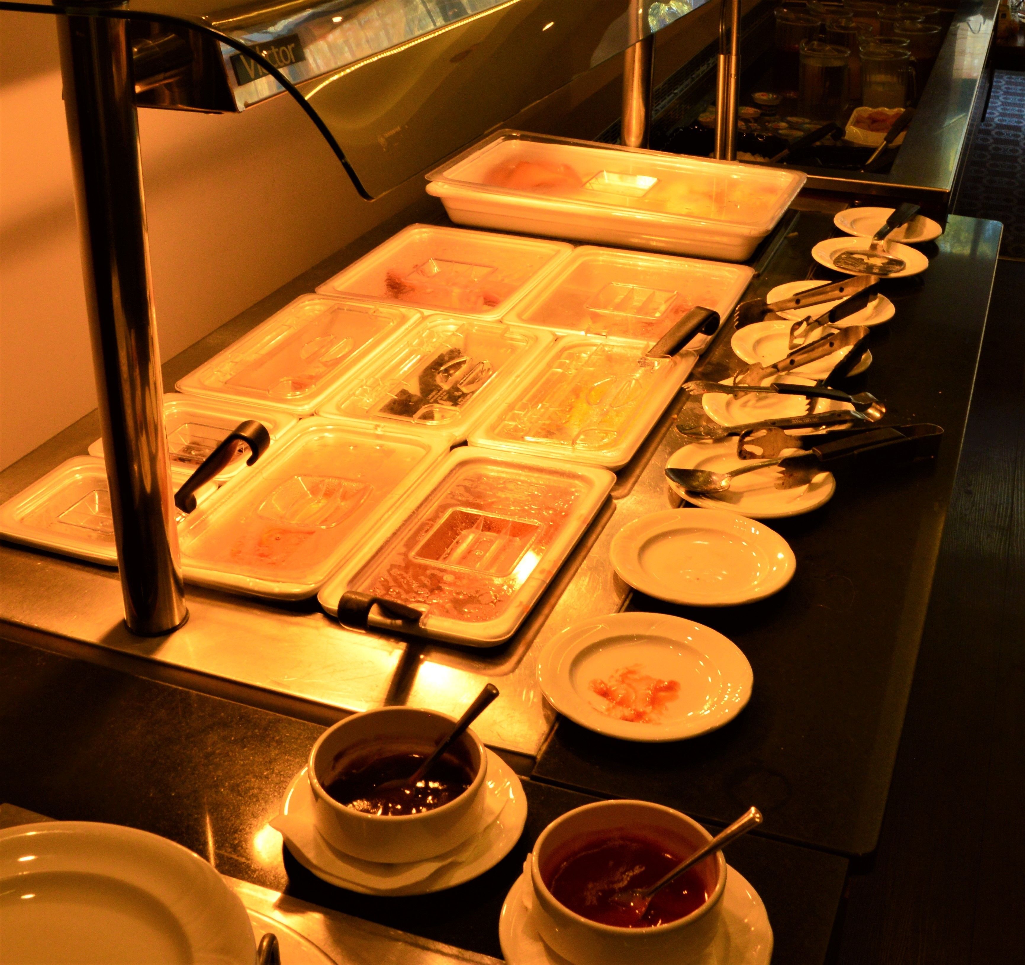Disposição de buffet com várias estações de serviço, incluindo bandejas e recipientes com comida, talheres e pratos.