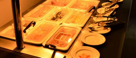 Disposição de buffet com várias estações de serviço, incluindo bandejas e recipientes com comida, talheres e pratos.
