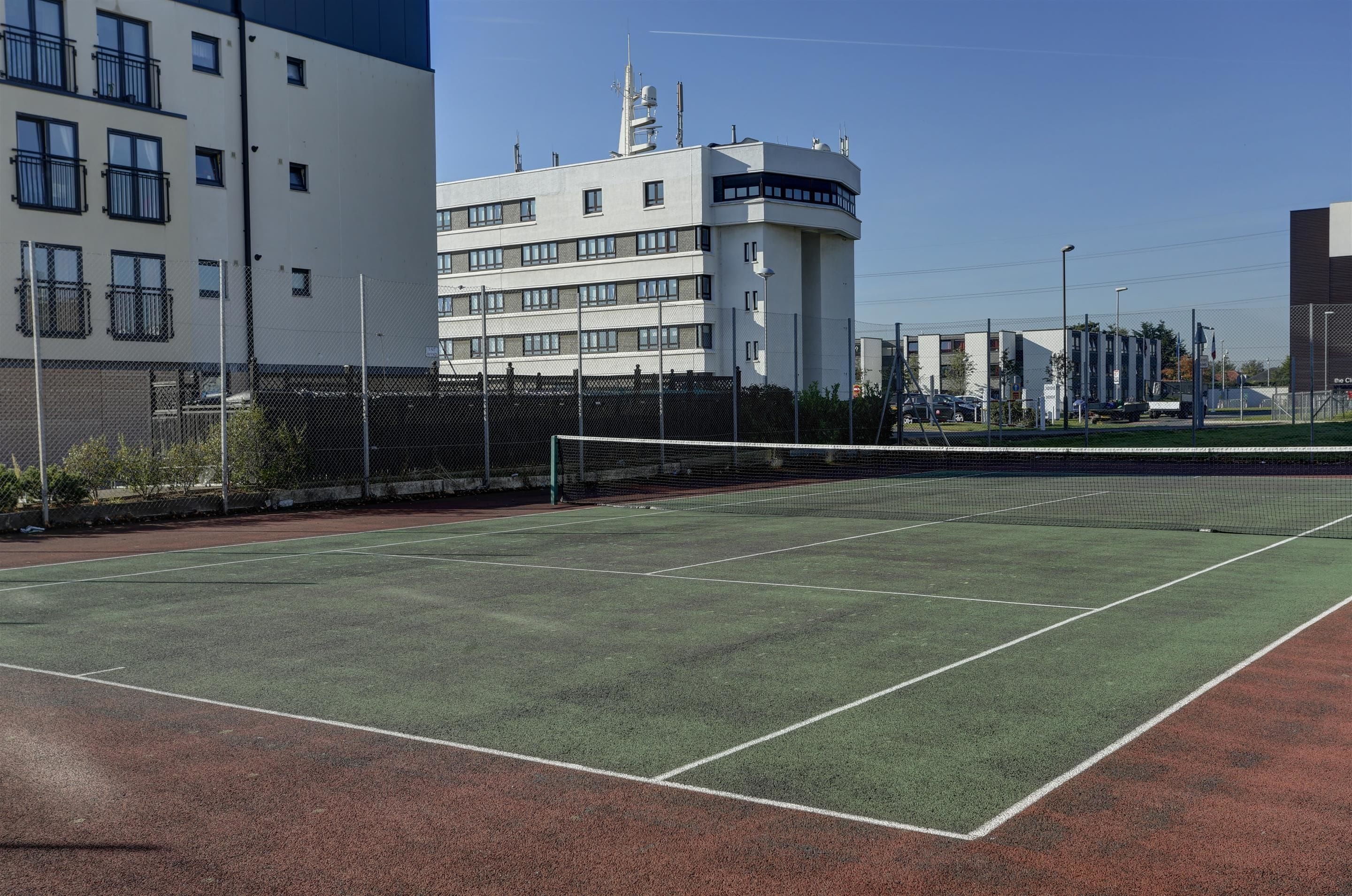 An outdoor tennis court with a net, surrounded by a fence, and buildings in the background.