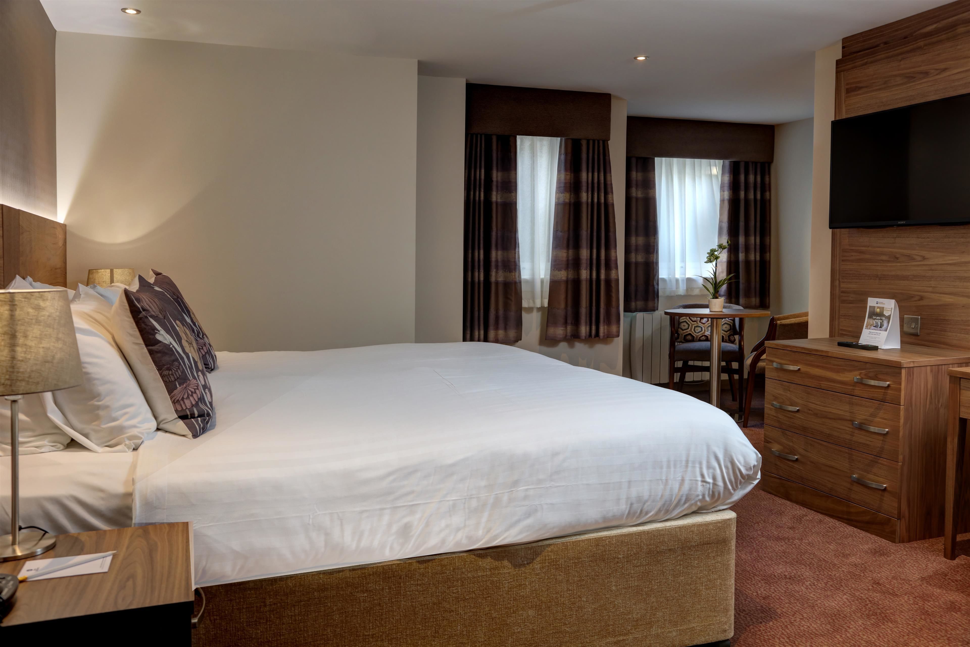 A hotel room with a large bed, a wooden dresser, a desk with a chair, and a flat-screen TV mounted on the wall.