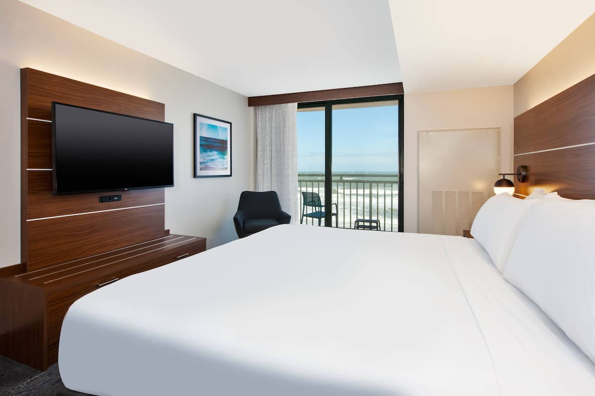 A modern hotel room with a large bed, a flat-screen TV mounted on a wooden panel, a small desk, a chair, and a balcony with a view of the beach.