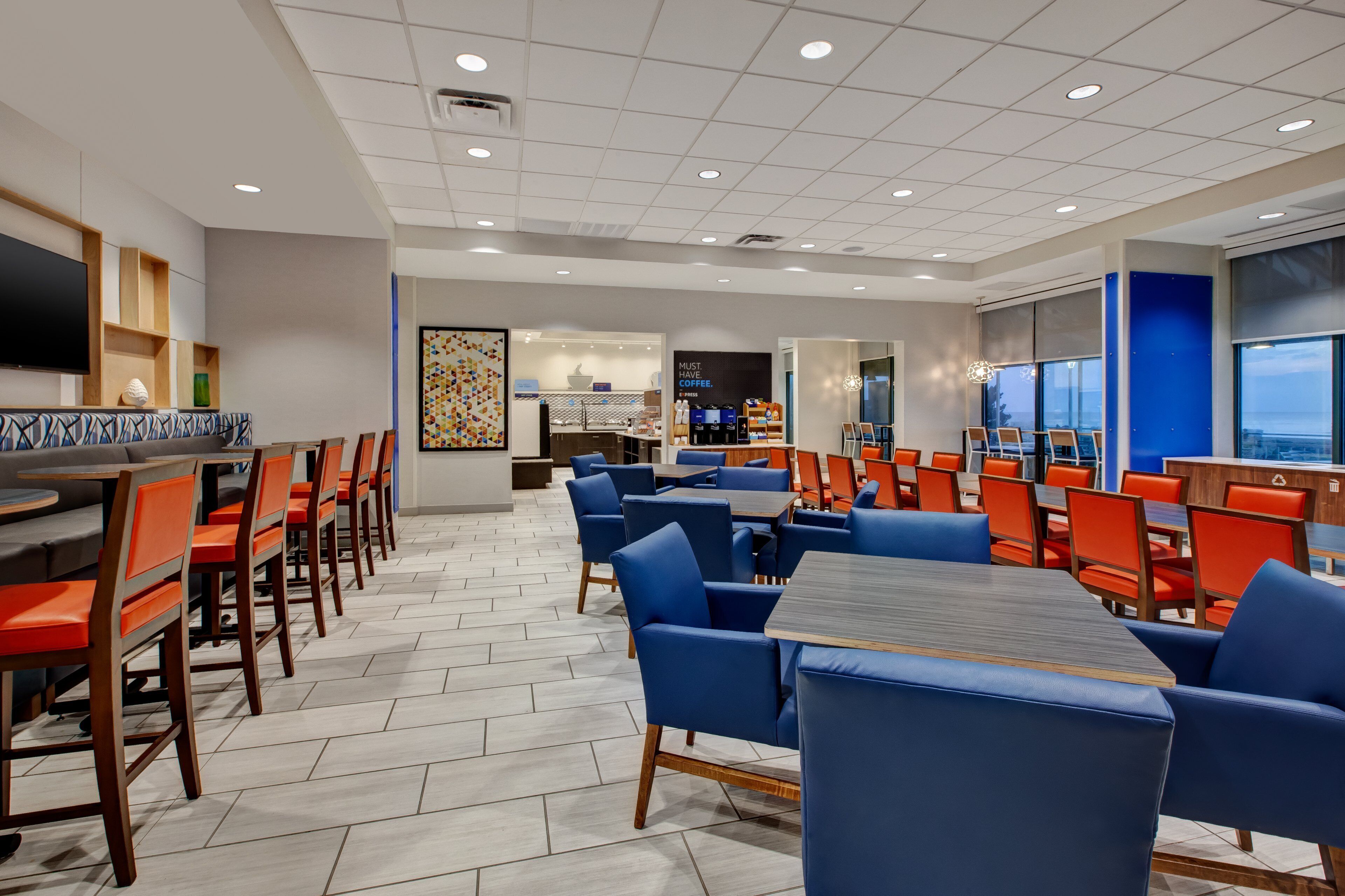 A modern dining area with multiple tables and chairs, a flat-screen TV mounted on the wall, and a large window offering a view.