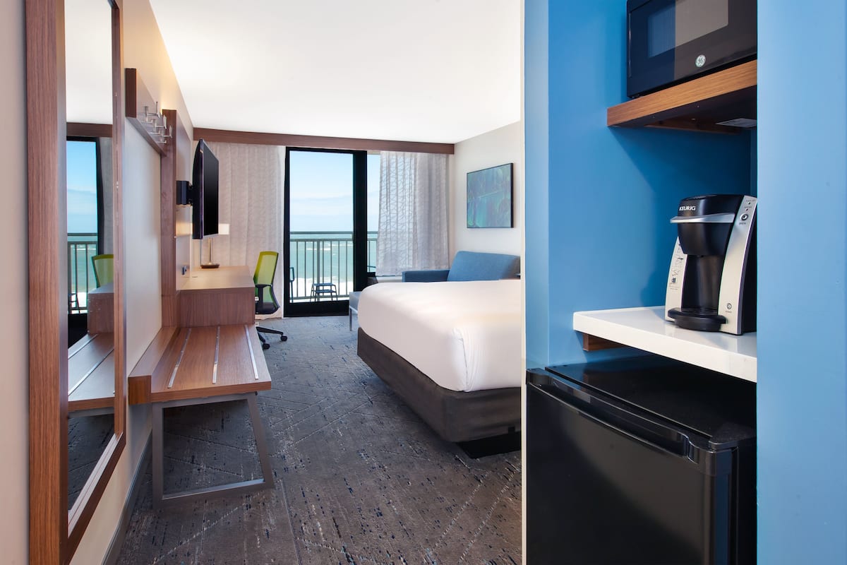 A hotel room with a bed, a desk, a chair, a coffee machine, and a view of the outdoors.