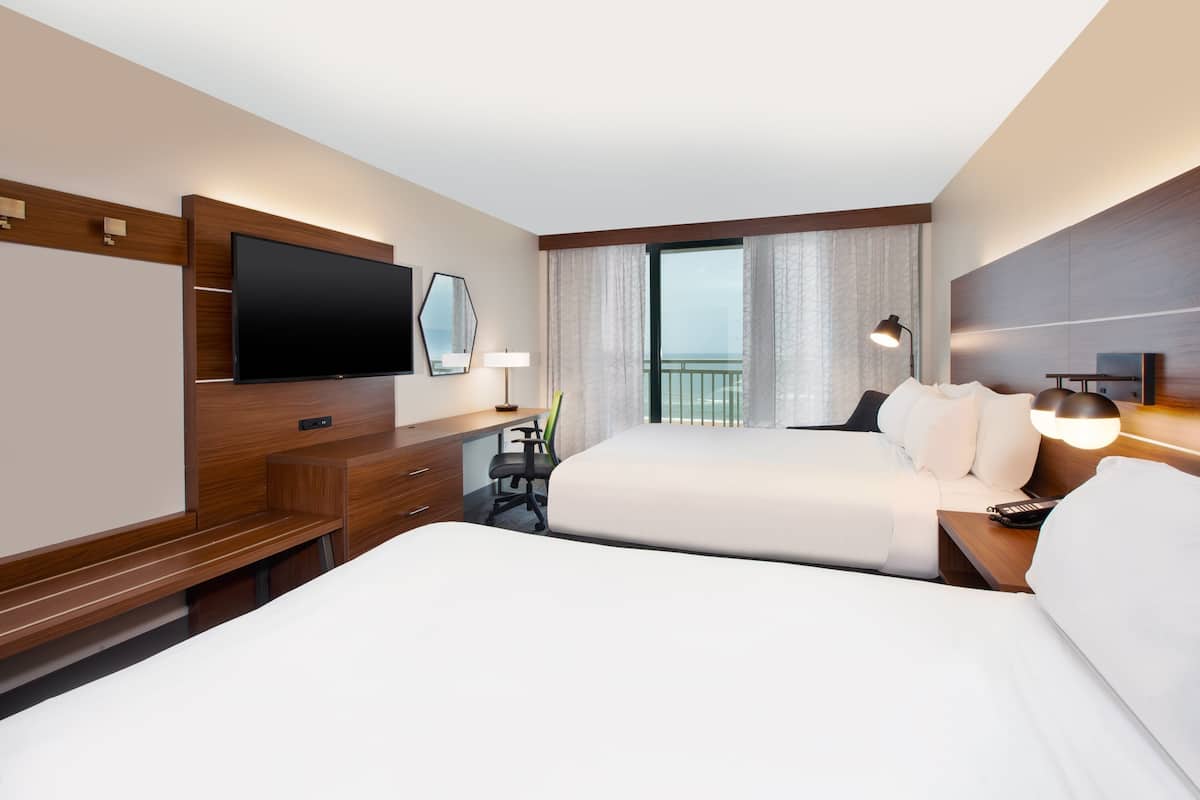 A hotel room with two beds, a desk, a flat-screen TV, a lamp, and a view of the sea.
