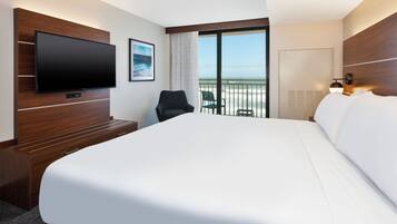 A hotel room with a large bed, a flat-screen TV, a desk, a chair, a balcony with a view of the sea, and a window with curtains.