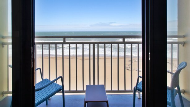 A balcony with two blue chairs and a small table, overlooking a beach with a railing and ocean view. - Holiday Inn Express & Suites VA Beach Oceanfront by IHG (Virginia Beach)