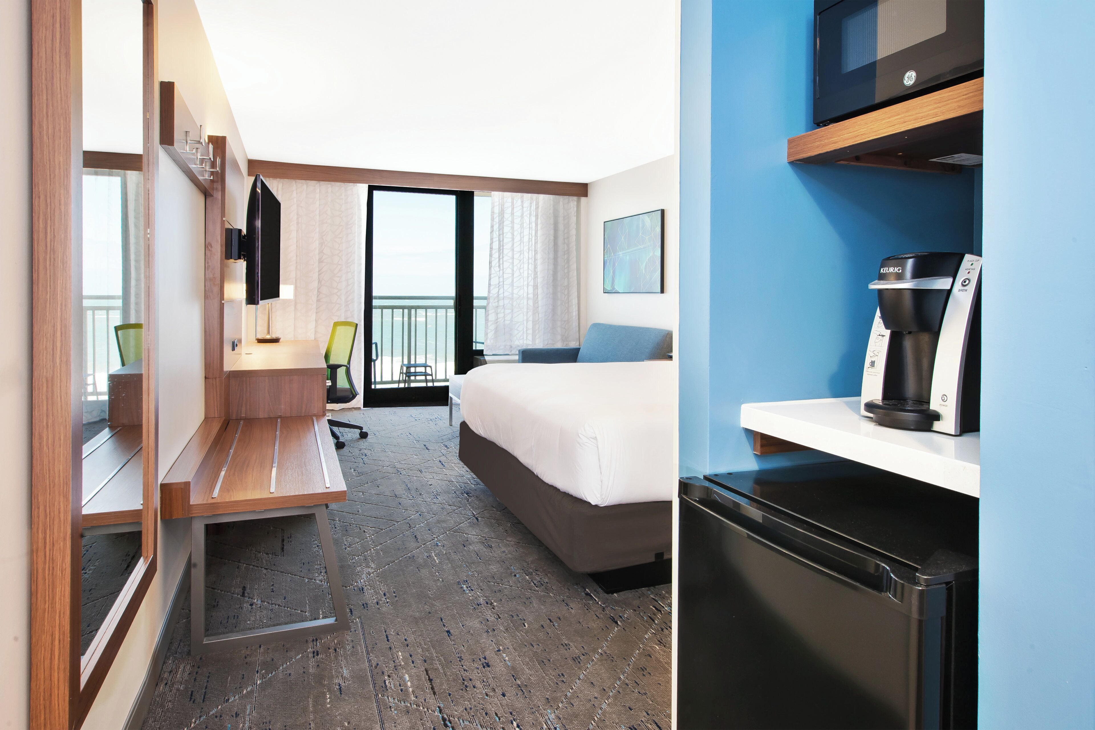 A hotel room with a bed, a desk, a chair, a television, a coffee machine, and a view of the sea.