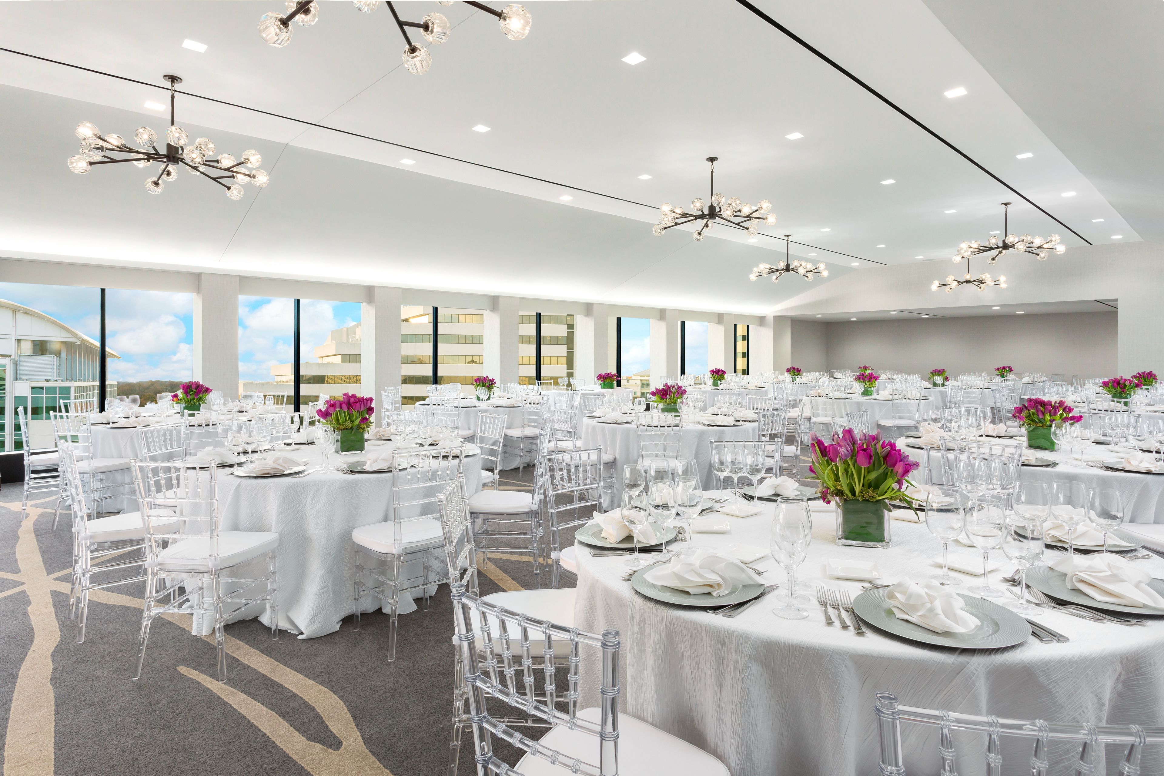 A large banquet hall with round tables set for dining, clear chairs, and decorative vases with flowers.
