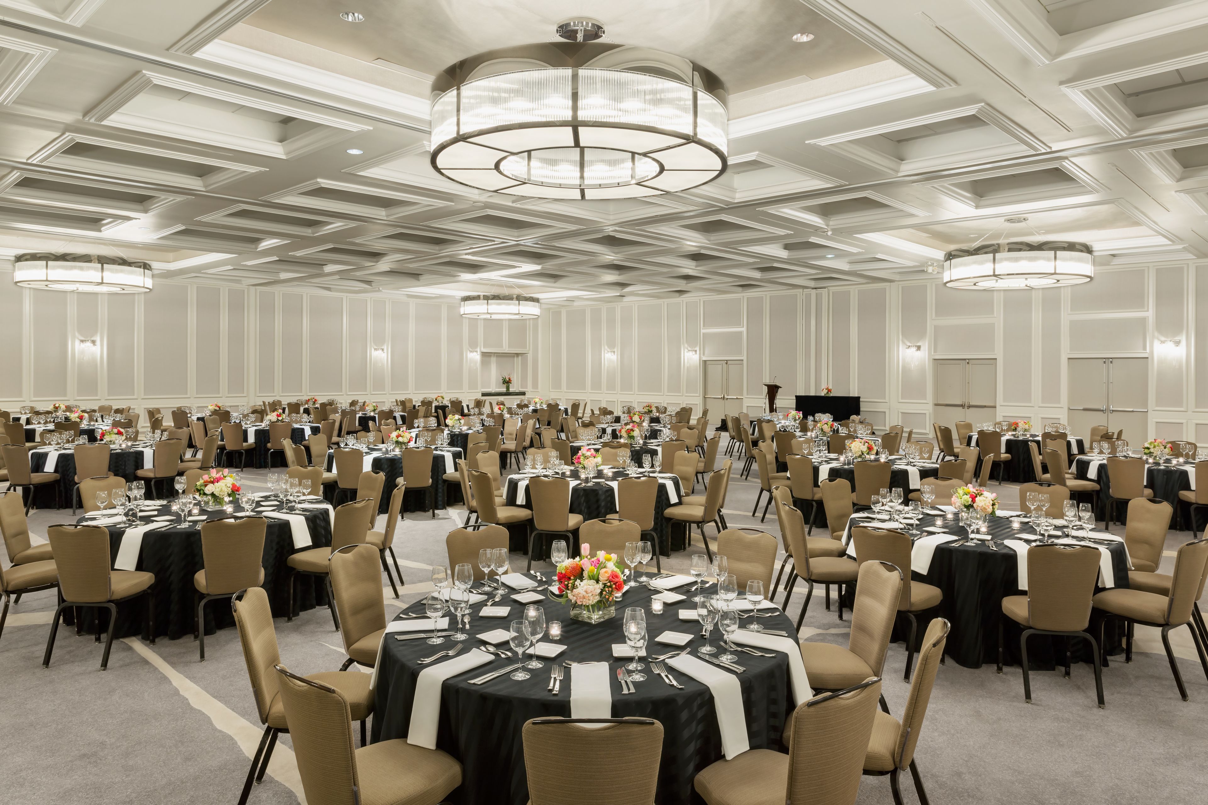 Un gran salón de banquetes con varias mesas redondas dispuestas para cenar, cada una con mantel negro y centros de mesa blancos.