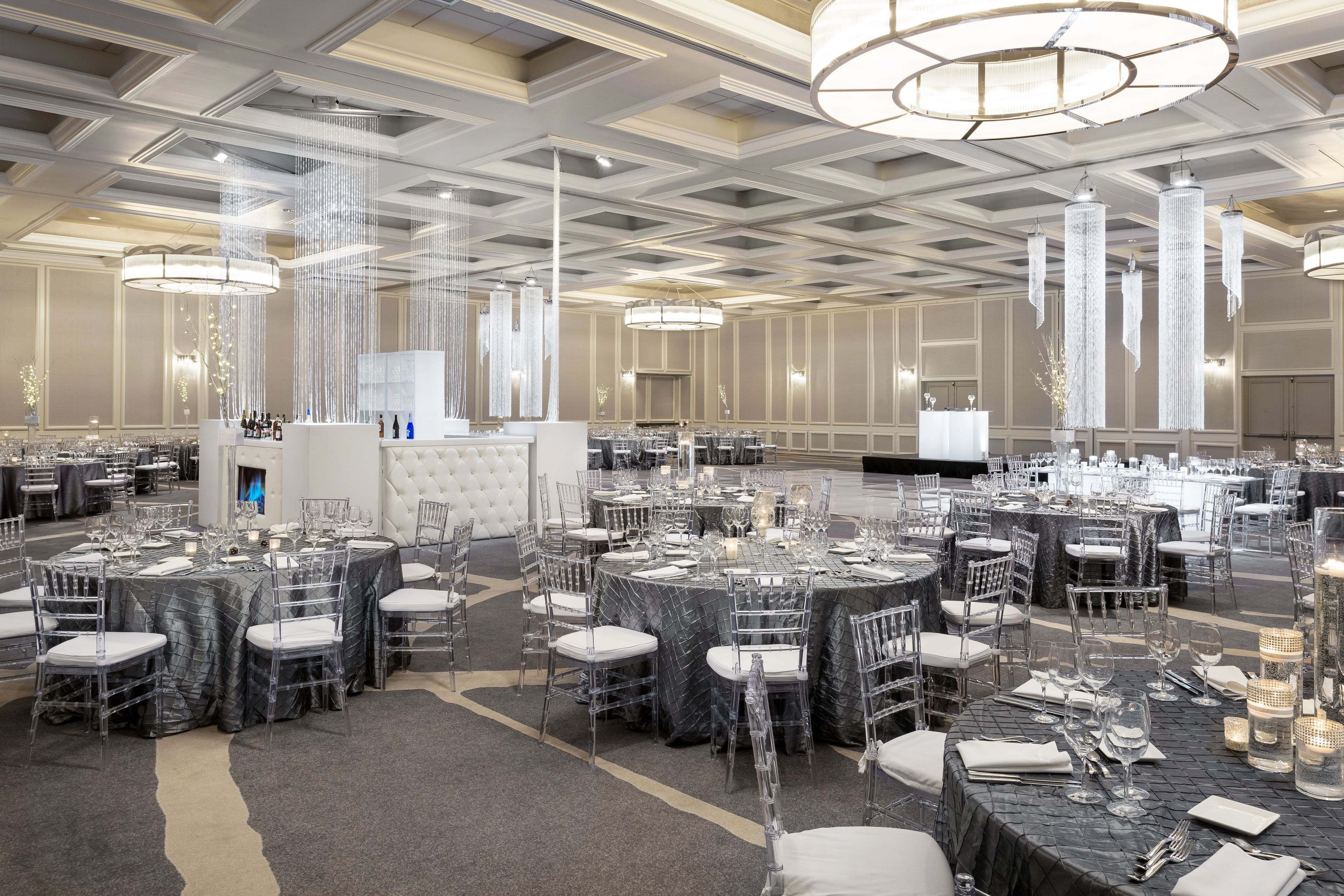 A banquet hall with round tables set for dining, clear chairs, and elegant chandeliers.