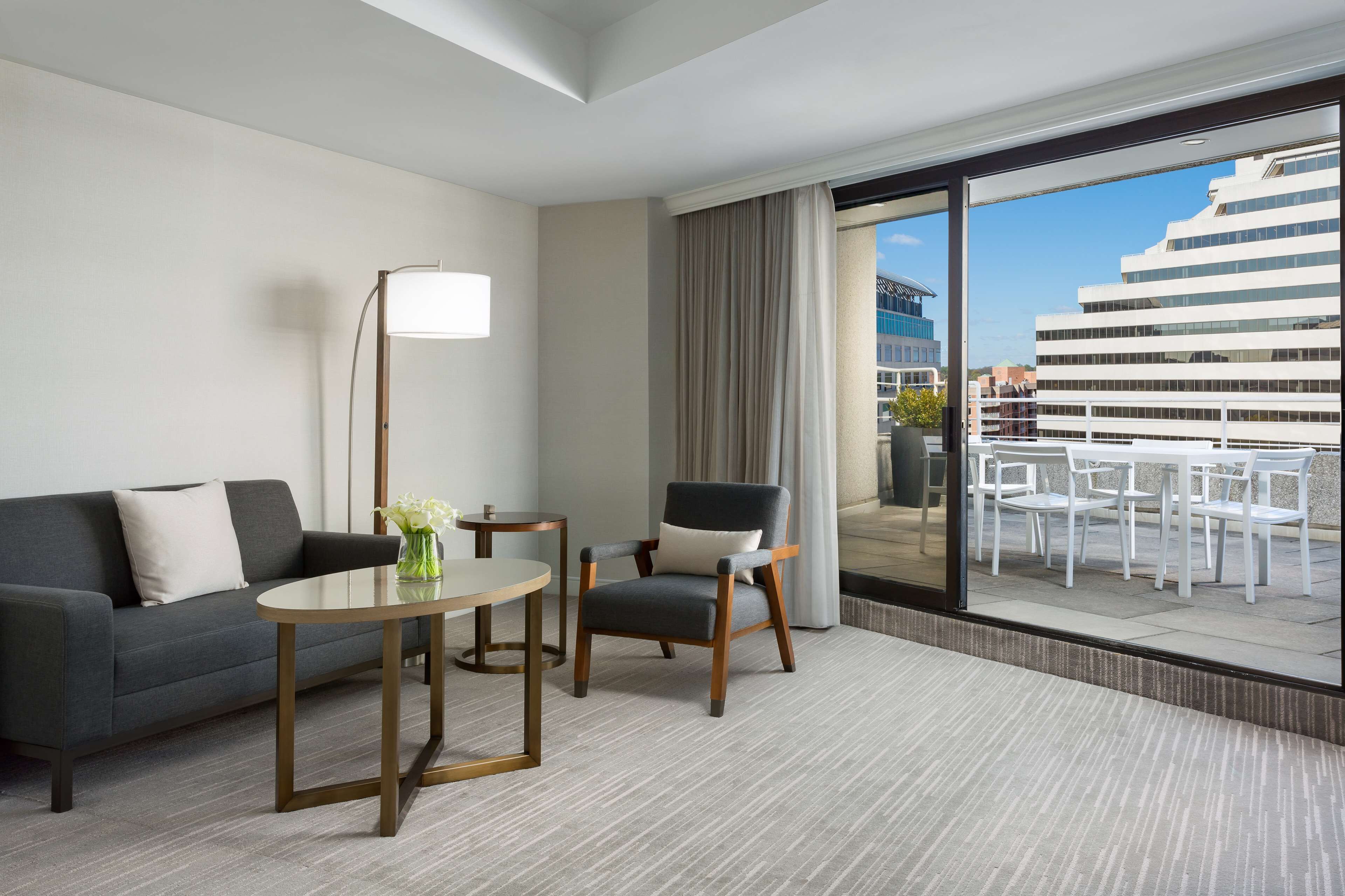 Quarto de hotel moderno com sofá cinzento, uma mesinha redonda, uma cadeira e uma grande janela com vista para um navio de cruzeiro e edifícios.