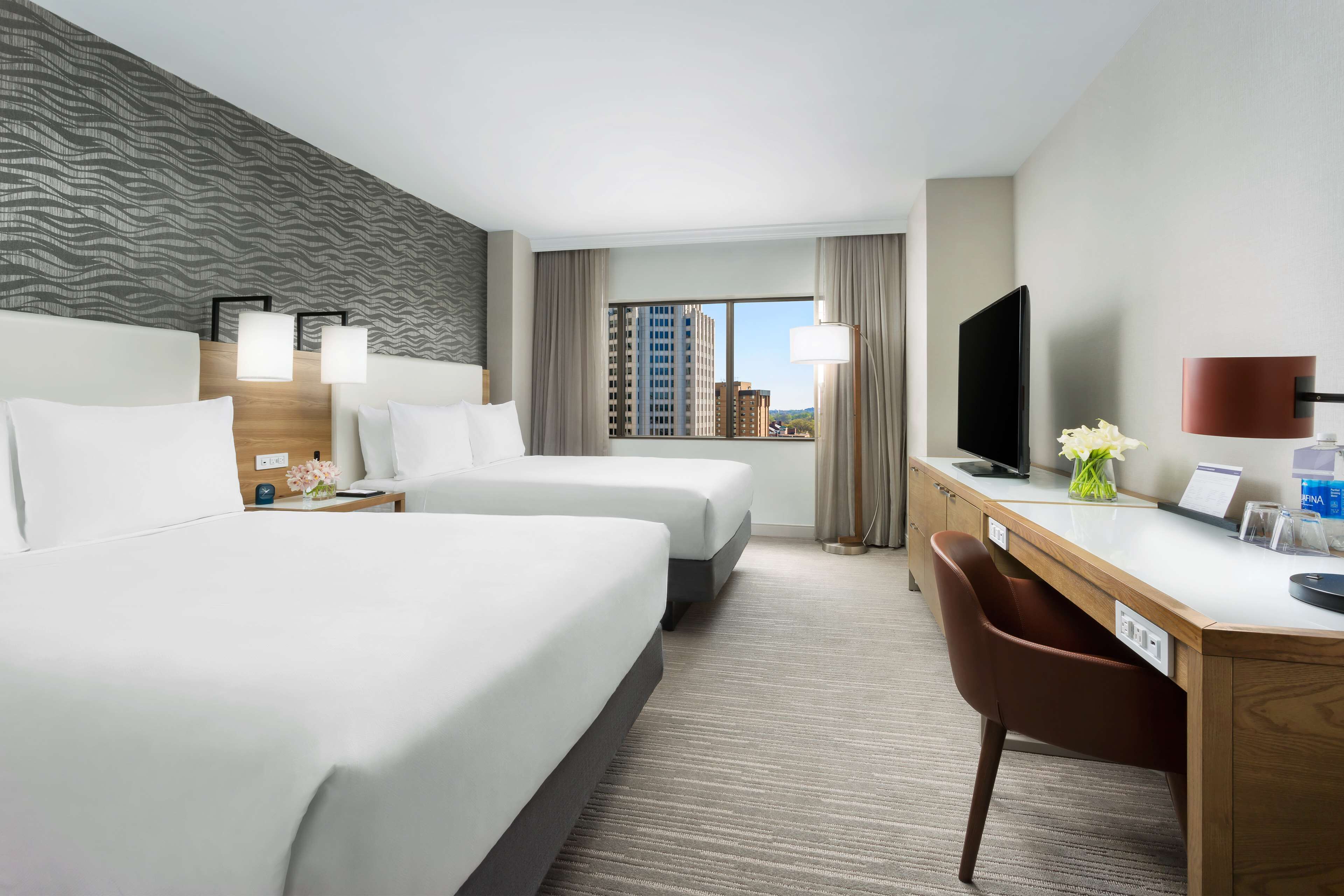 Habitación de hotel con dos camas, un escritorio, una silla, un televisor y una ventana con vistas a la ciudad.