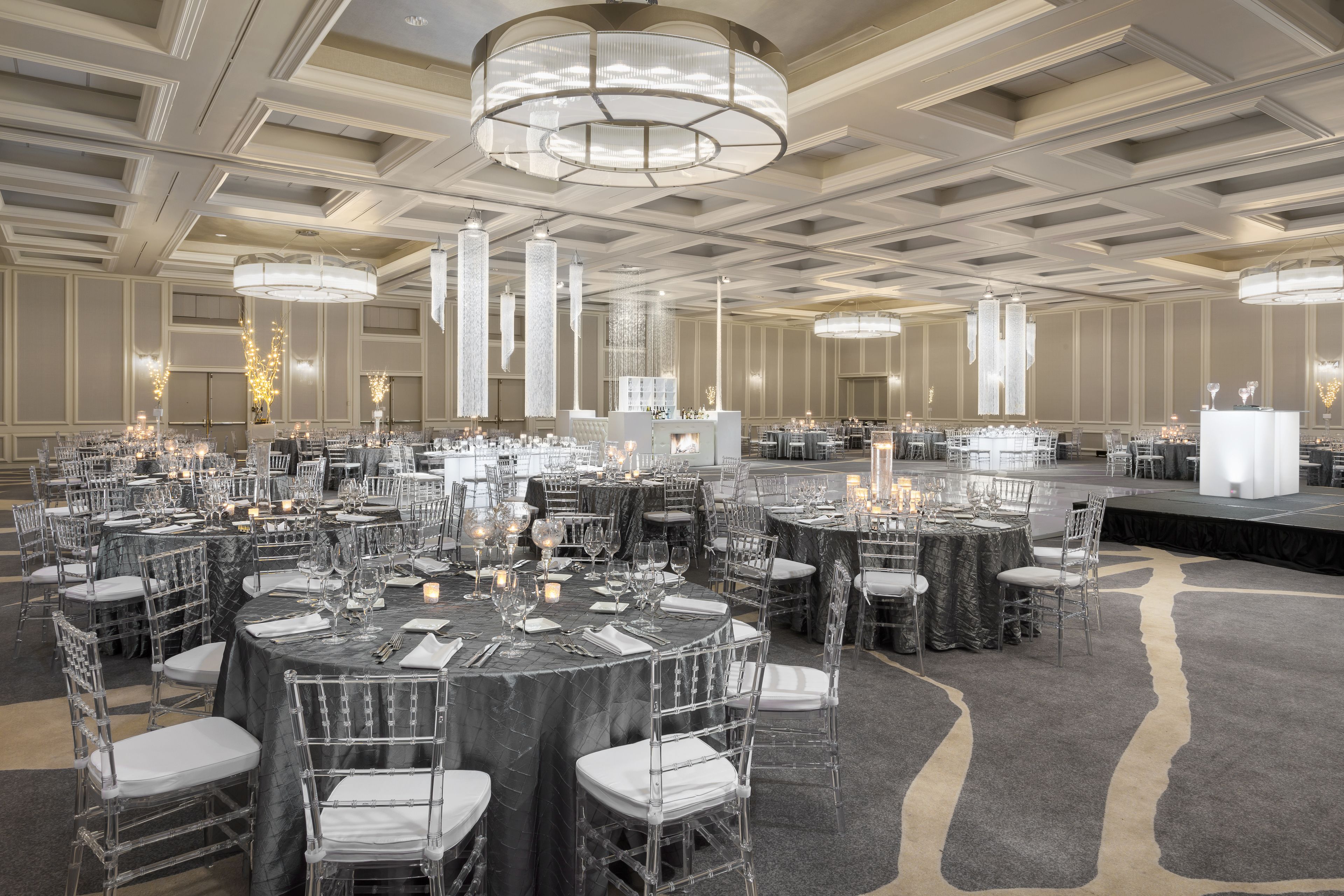 A banquet hall with round tables, clear chairs, and chandeliers.