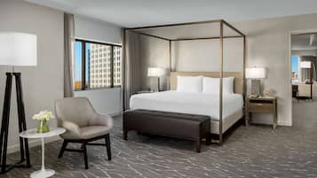 A modern hotel room with a four-poster bed, a round side table, a chair, and a small table with a vase of flowers.
