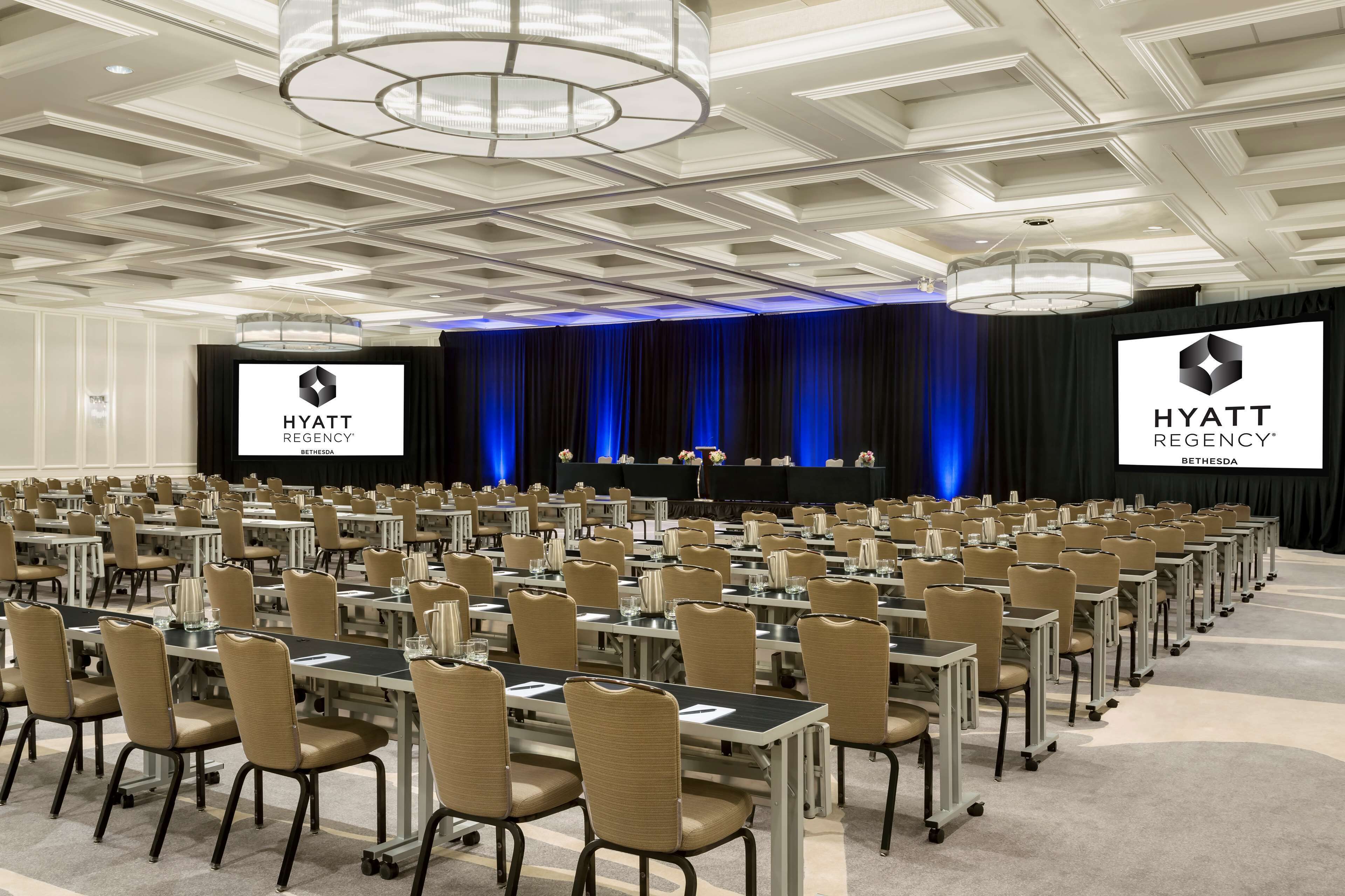 Un salón de conferencias con filas de mesas y sillas, un escenario con telón de fondo azul y una pantalla grande que muestra el logotipo de Hyatt Regency.