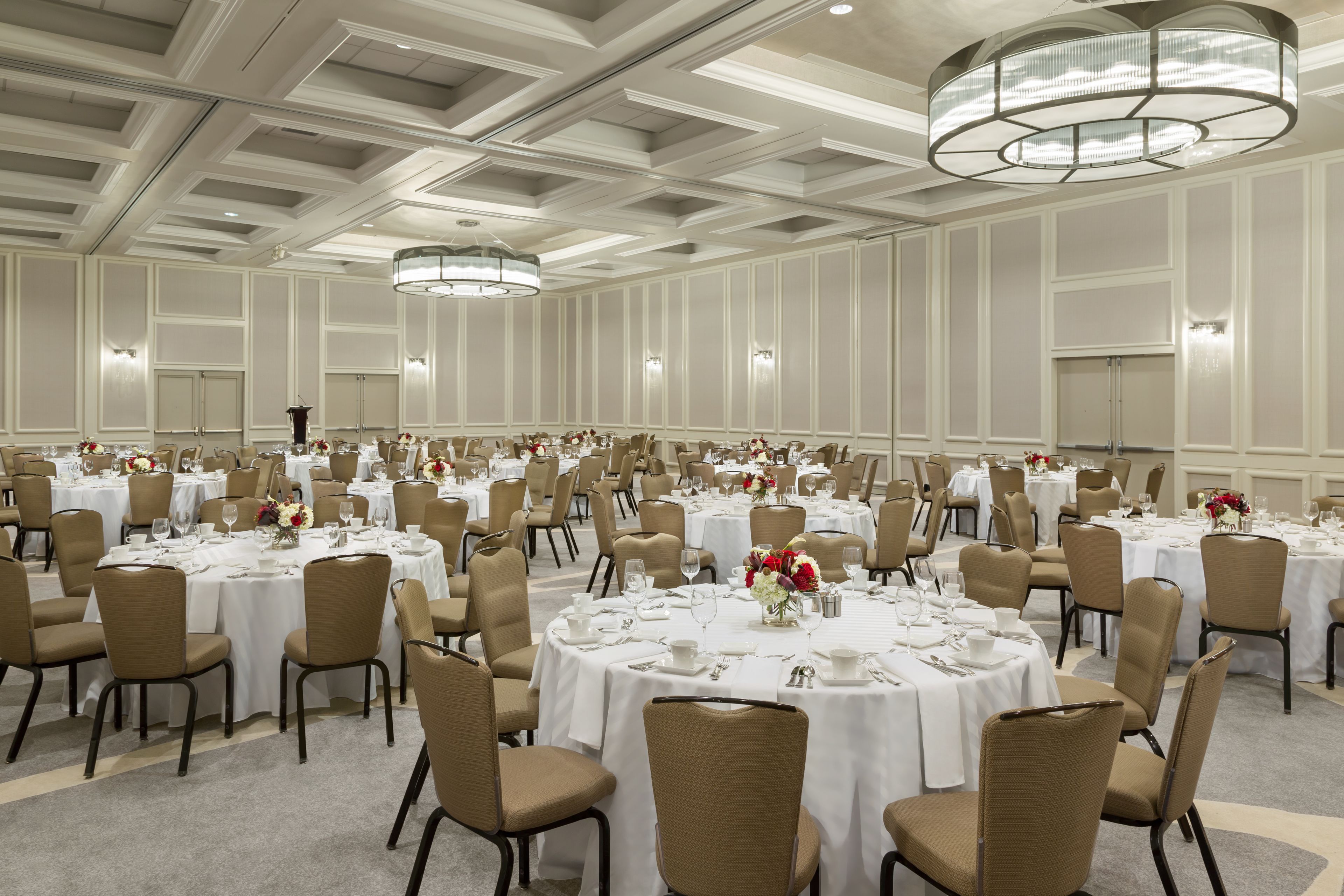 Un amplio salón de banquetes con varias mesas redondas dispuestas para comer, cada una con manteles blancos y sillas color beige.