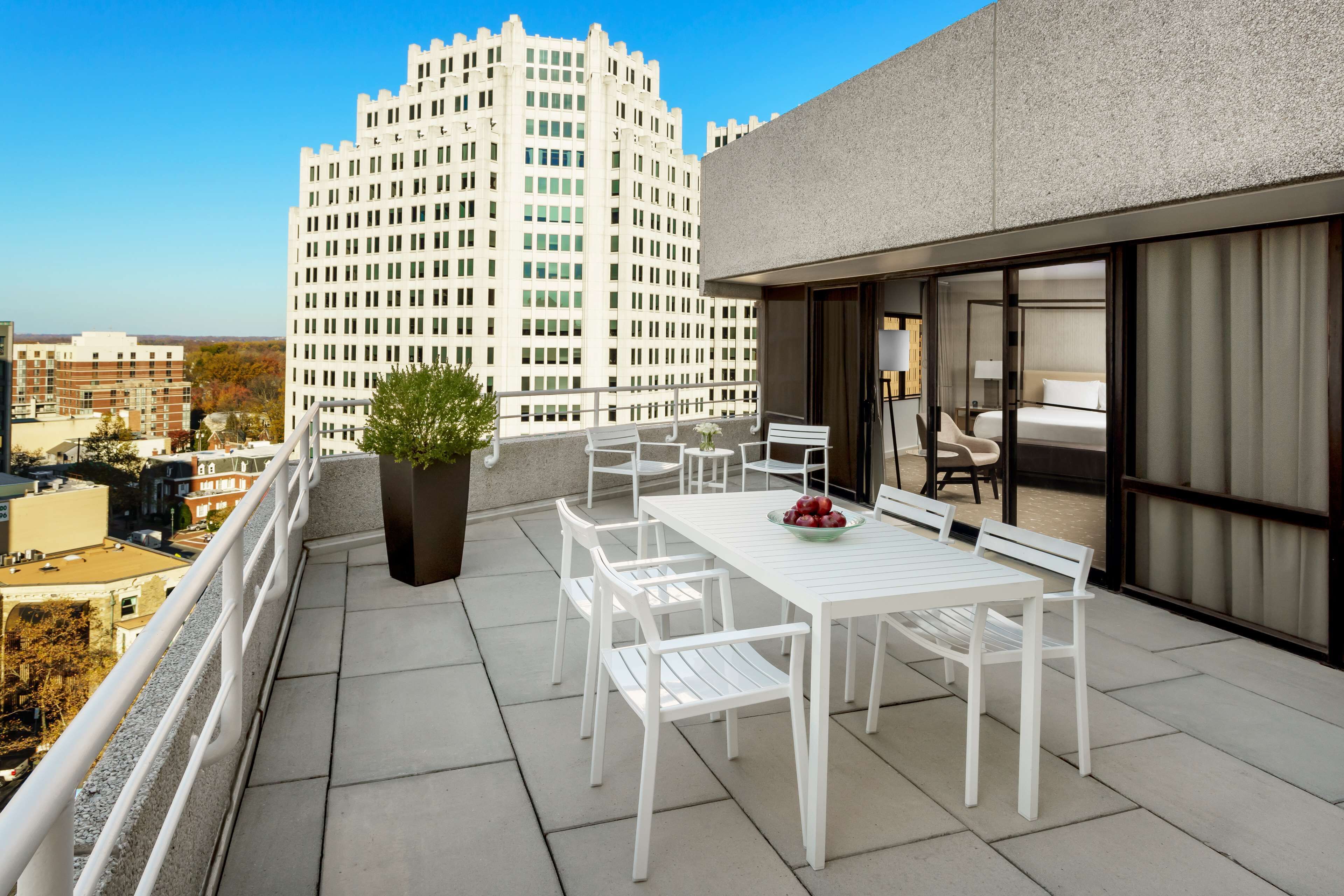 A rooftop terrace with a white table and chairs, a potted plant, and a view of a tall building in the background.
